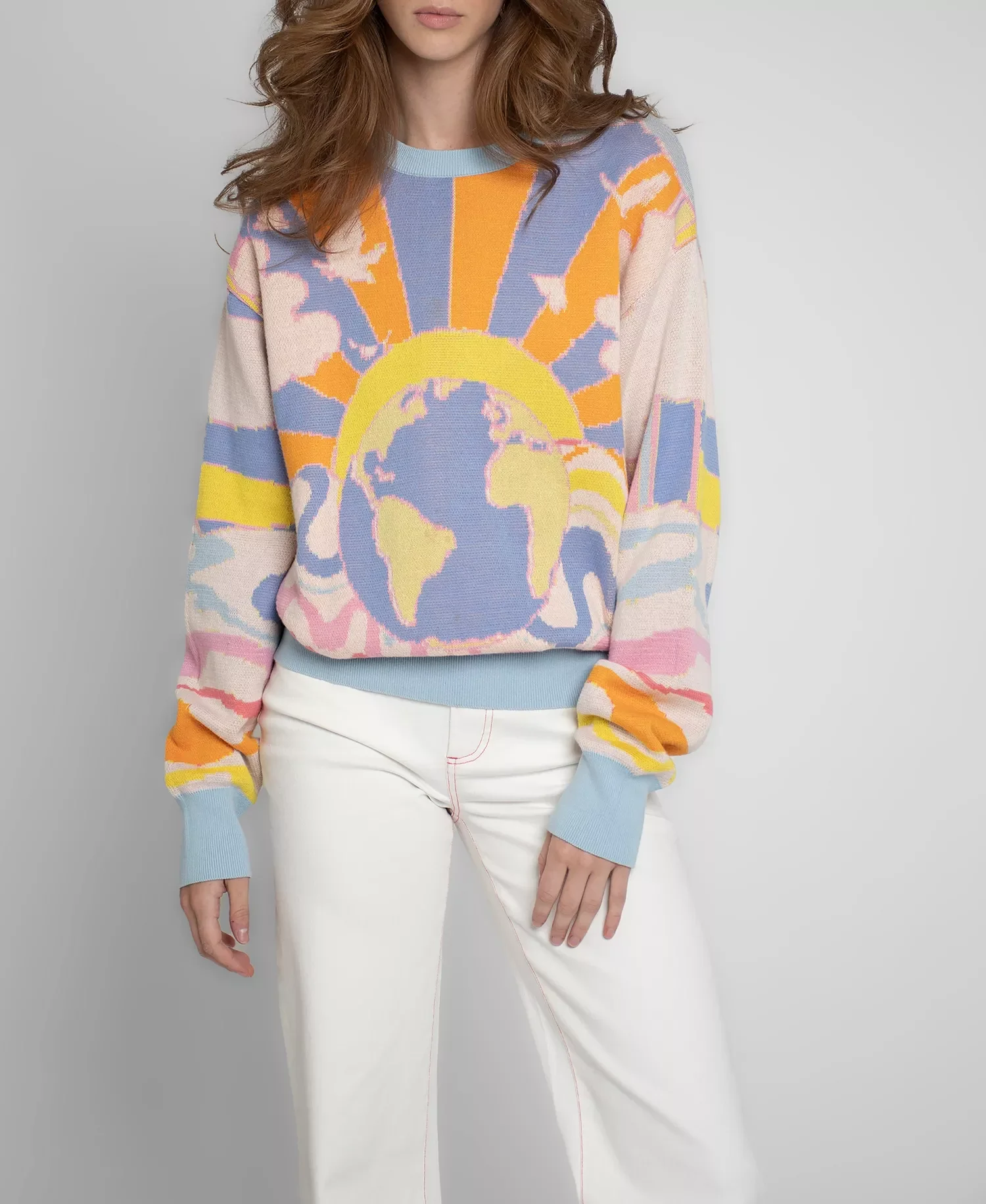 A woman wearing a colorful sweater with a sun and earth design and white pants.