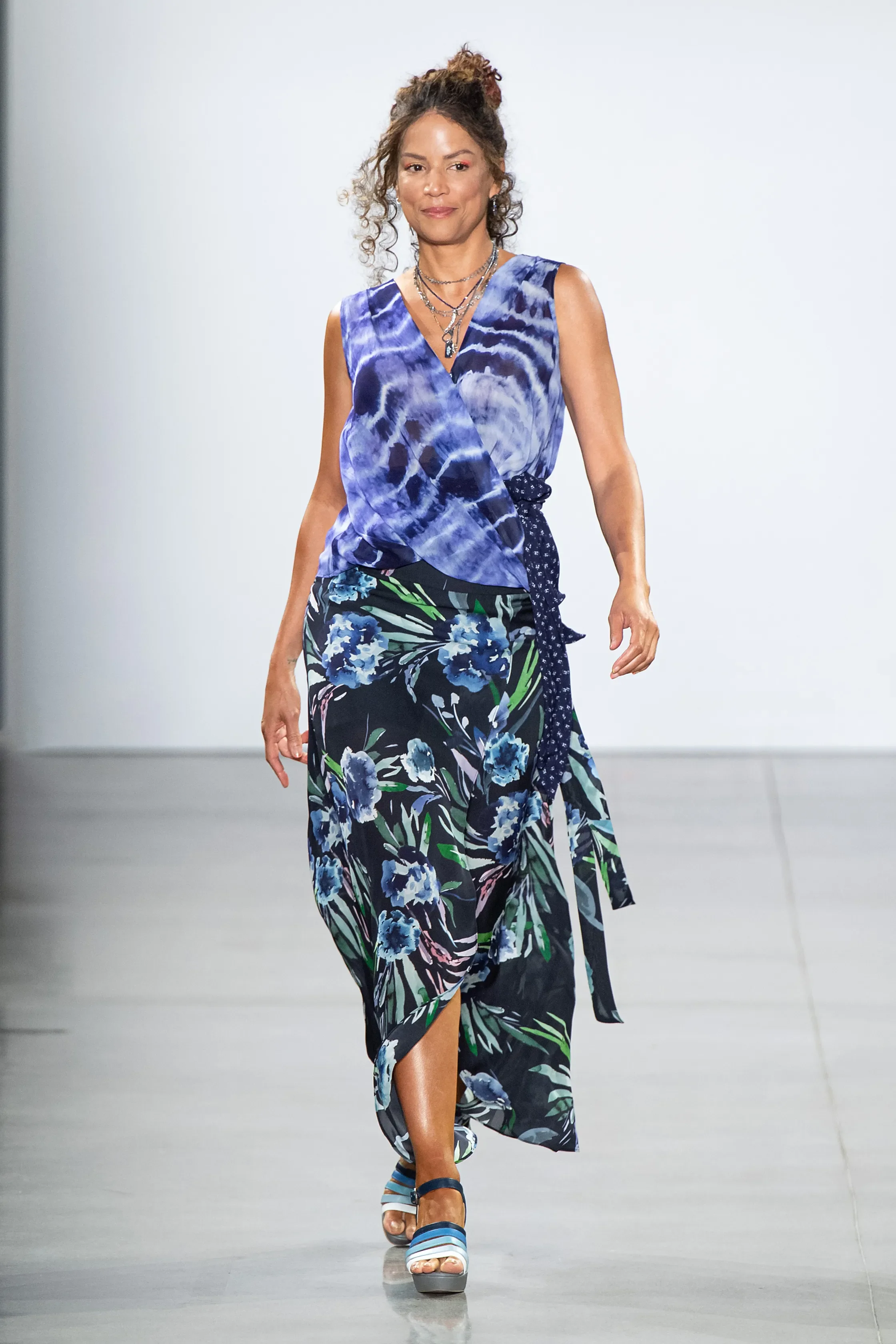 A woman walking on a runway wearing a blue tie-dye sleeveless top, a floral skirt with blue blossoms, and platform sandals, accessorized with layered necklaces.