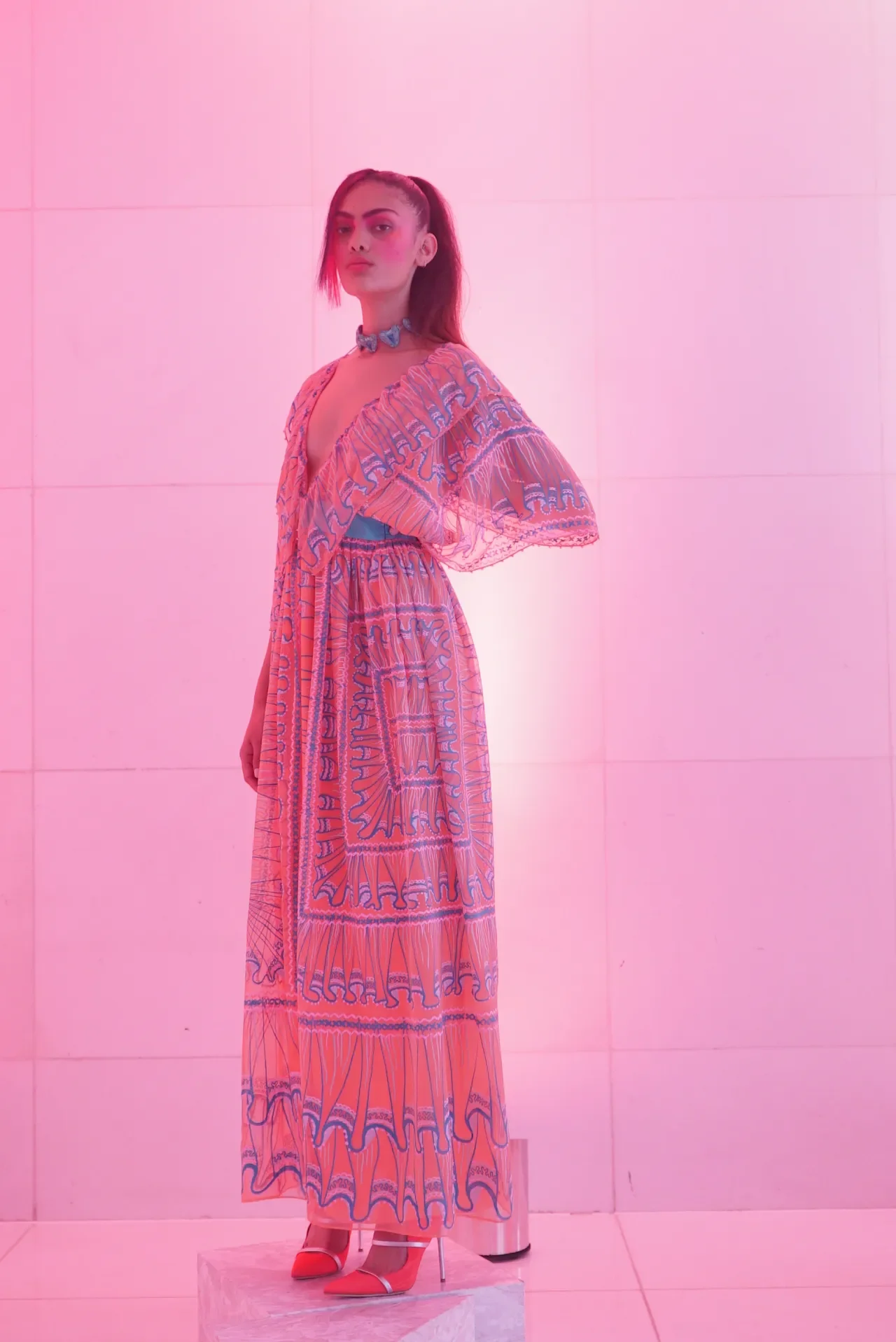 A woman wearing a pink patterned dress and red heels standing on a block against a pink wall illuminated with pink lighting.