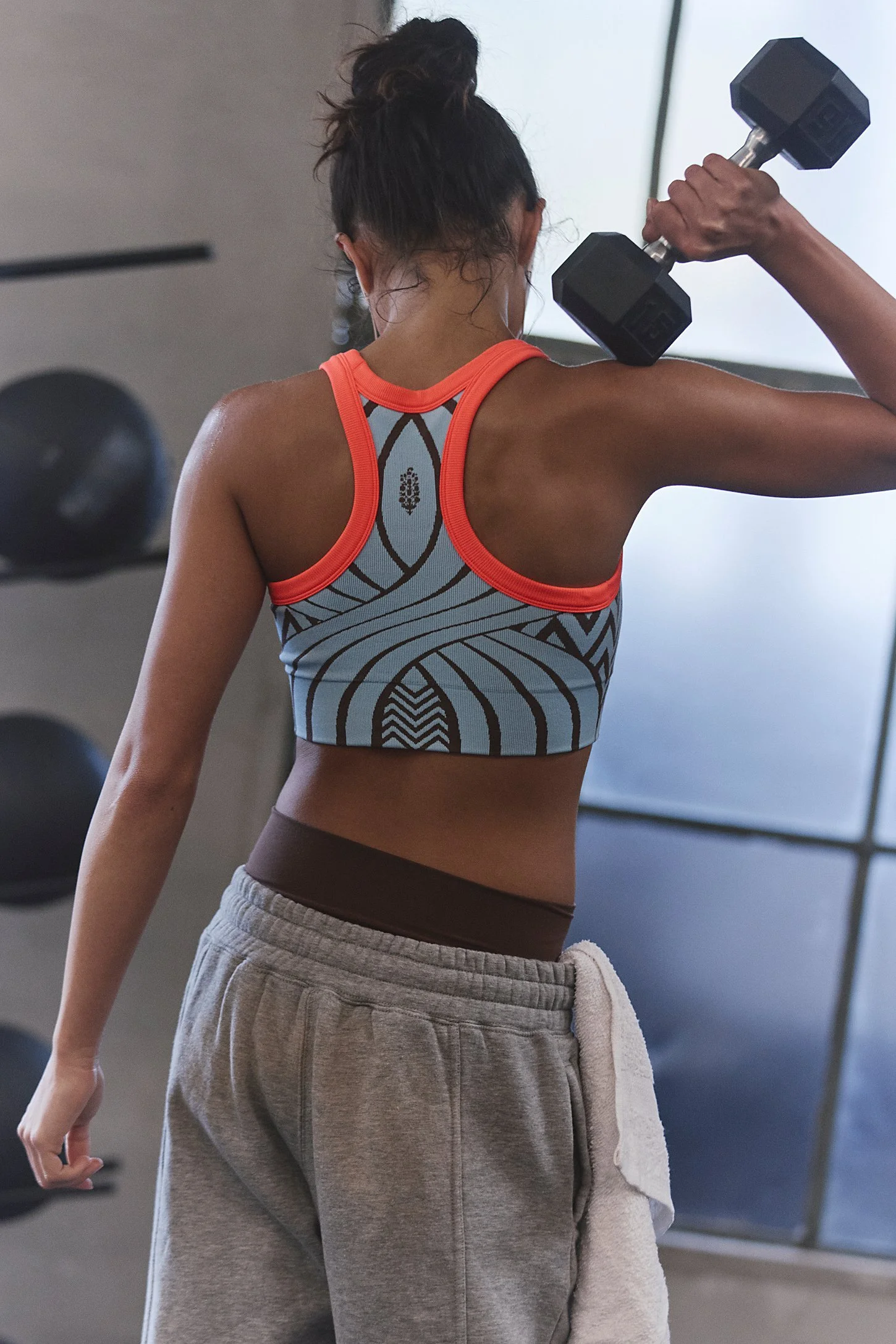 A woman with dark hair tied up in a messy bun, wearing a colorful athletic sports bra and gray sweatpants, lifting a dumbbell in a gym.