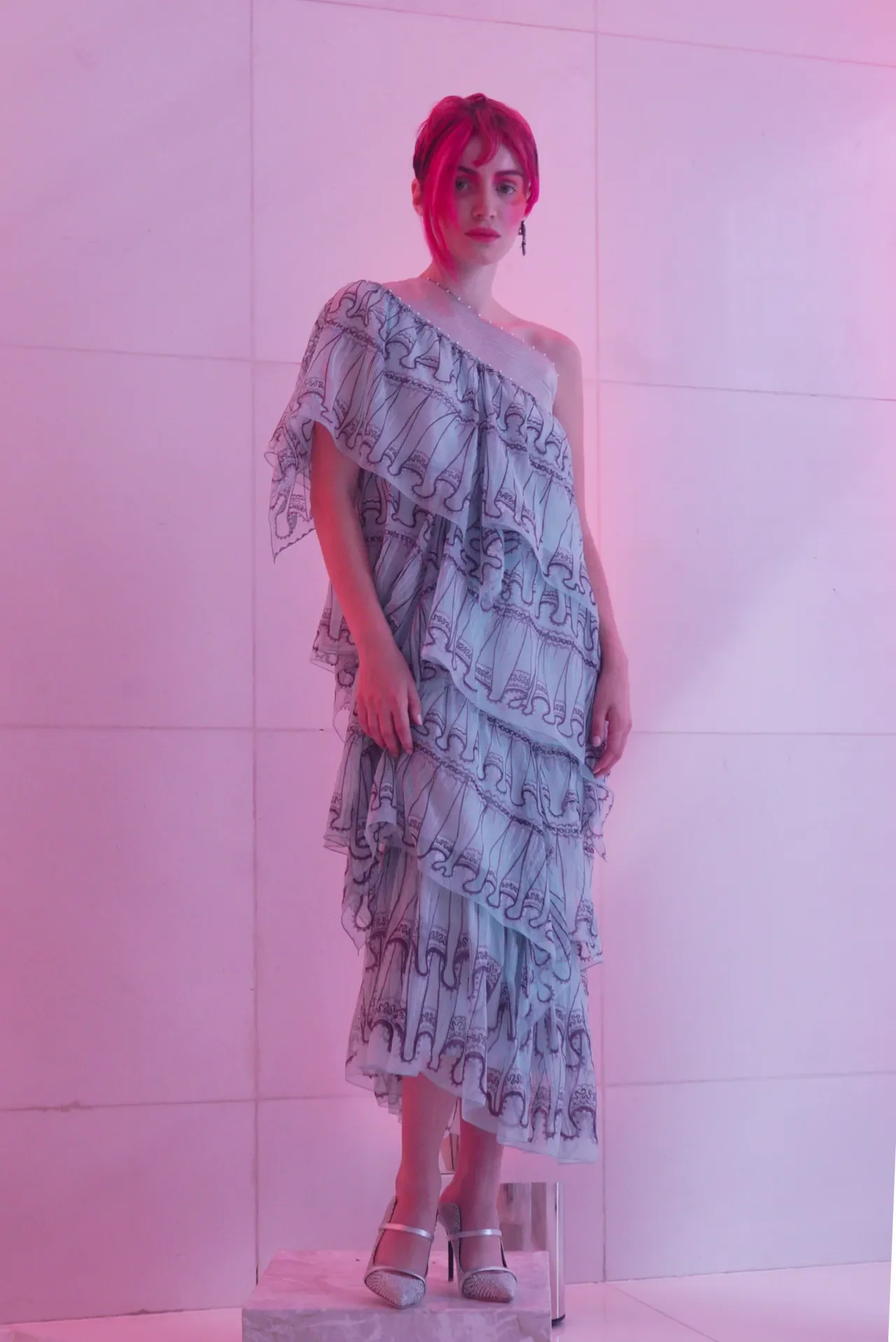 A woman with red hair wearing a layered, ruffled dress with a pattern, standing against a pink-lit wall, wearing high heels.