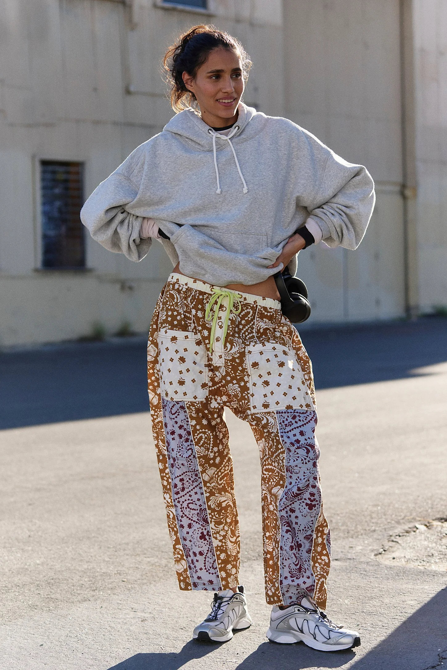 A woman wearing a light gray hoodie, patterned brown and white pants, and white sneakers standing outdoors on a paved surface with a beige building in the background.