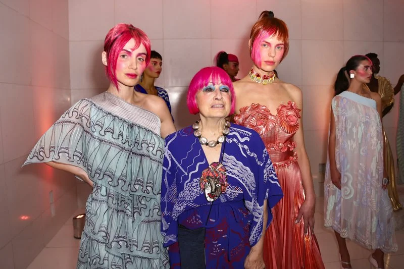 Three women with pink hair posing at a fashion event, dressed in colorful, artistic outfits, with several other models in the background.