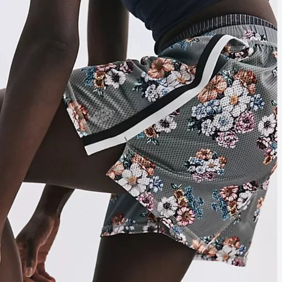 Close-up of a person wearing gray shorts with a floral pattern and a white and black stripe, sitting with one knee up and one hand resting on the ground.