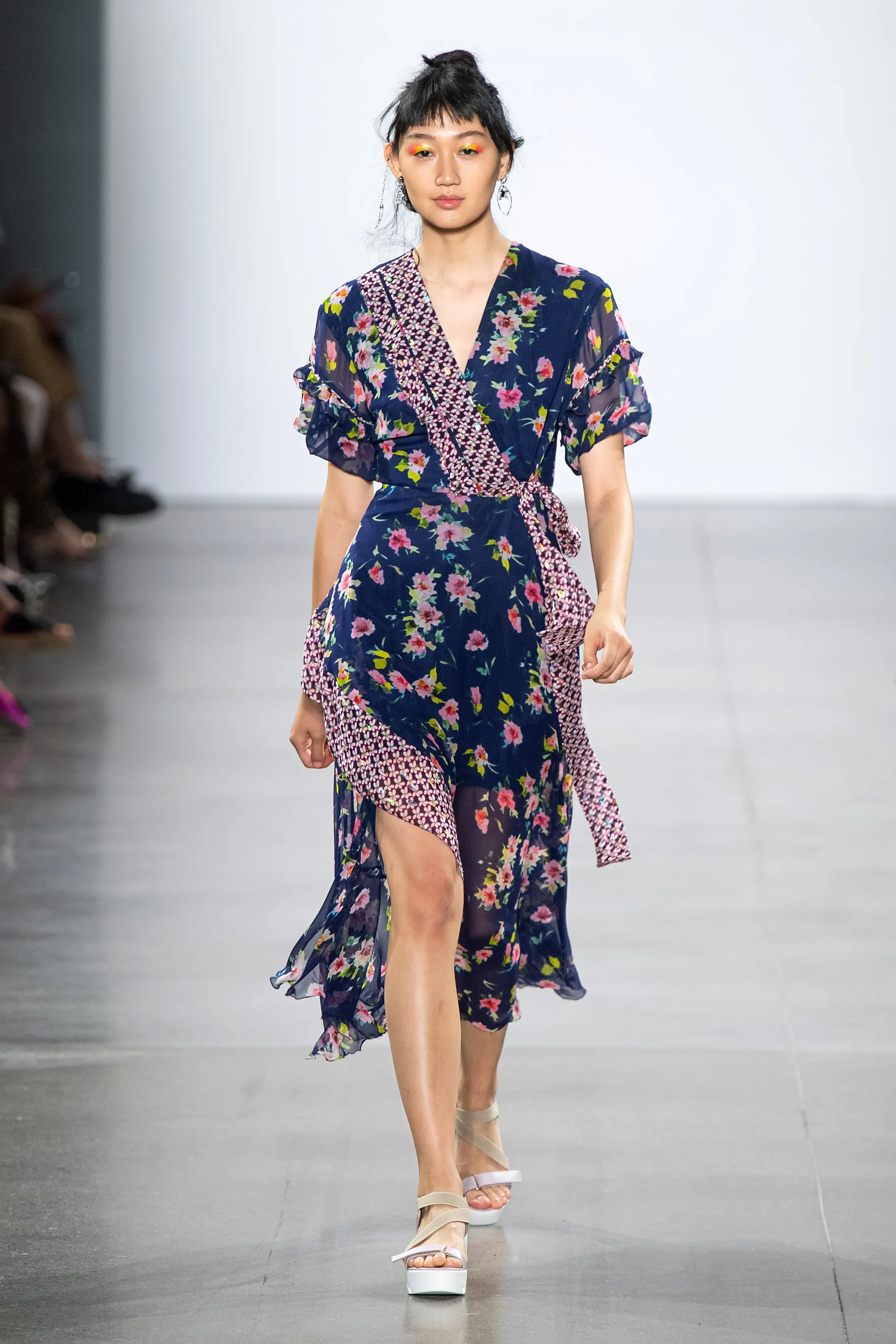 Model walking on runway in a navy blue floral wrap dress with ruffled sleeves and side slit, paired with beige platform sandals.