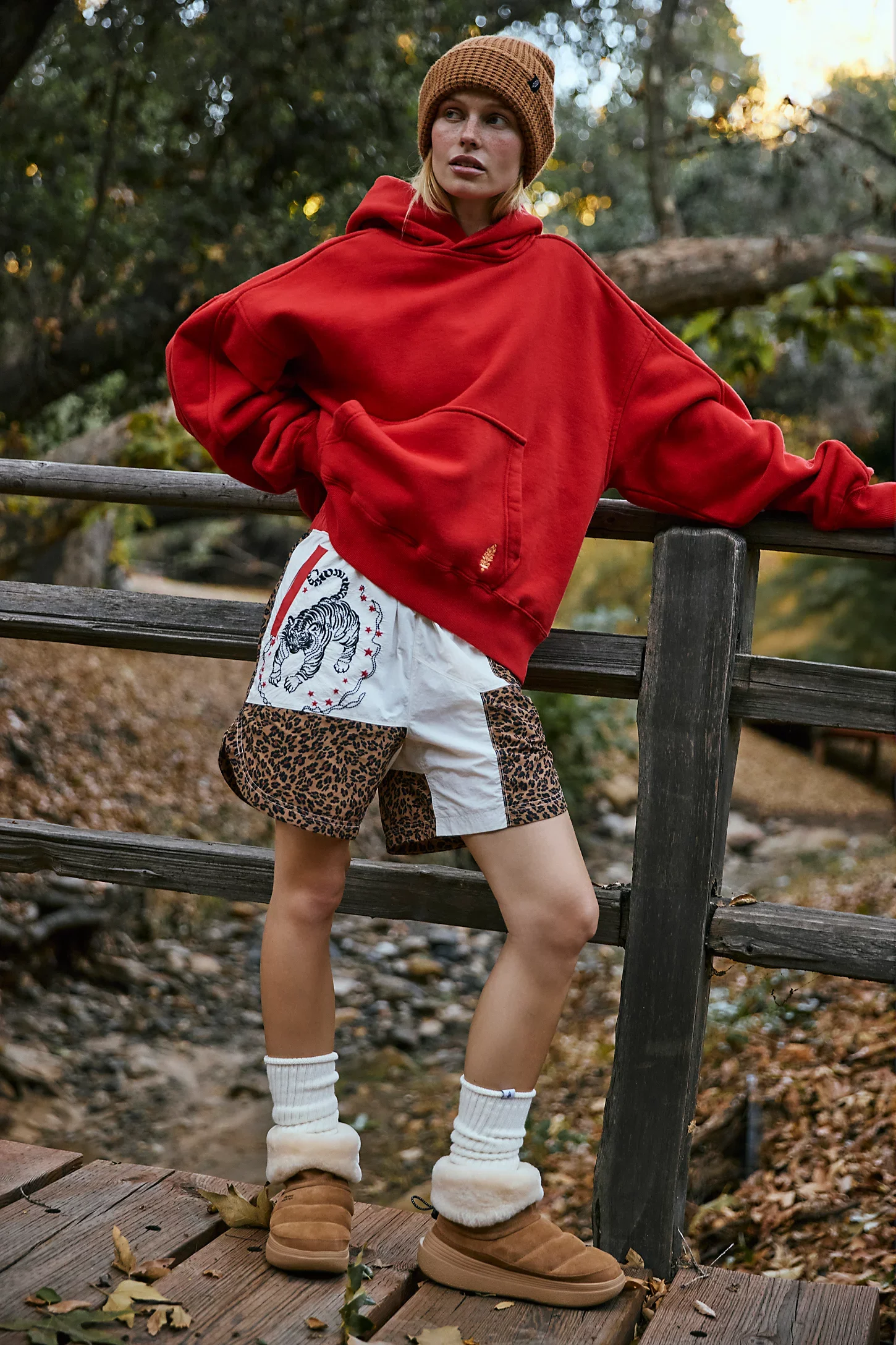 A woman wearing a red hoodie, brown beanie, leopard print shorts, white socks, and tan shoes stands outdoors on a wooden bridge with trees and fallen leaves in the background.