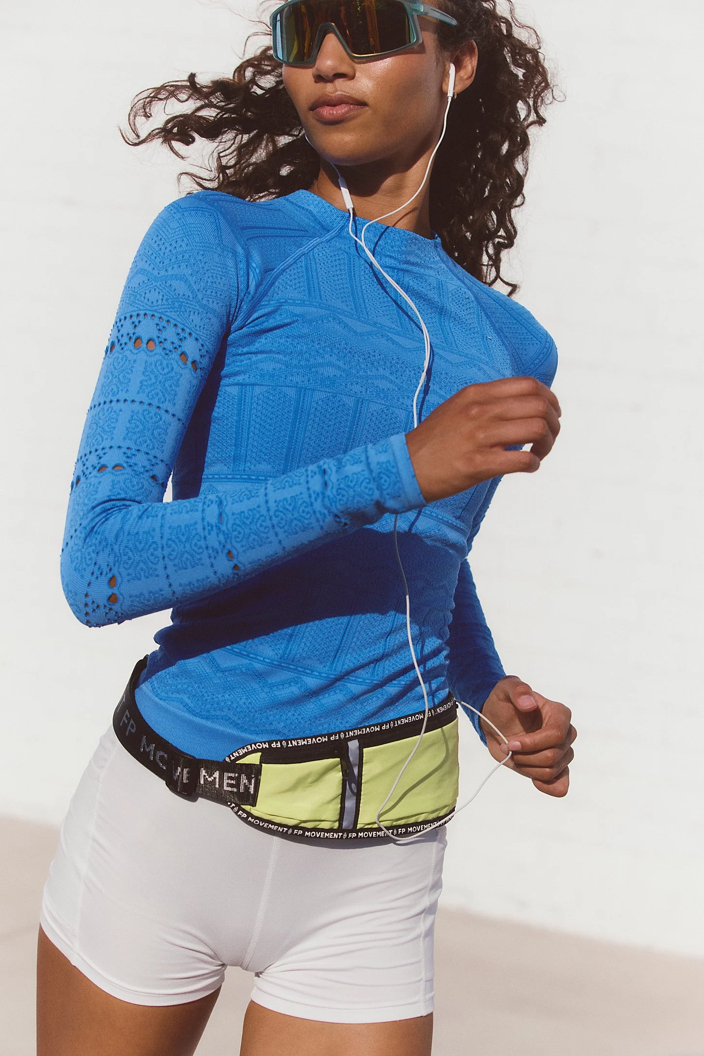 A woman running outdoors, wearing reflective sunglasses, earphones, a blue long-sleeve athletic top, and white shorts.