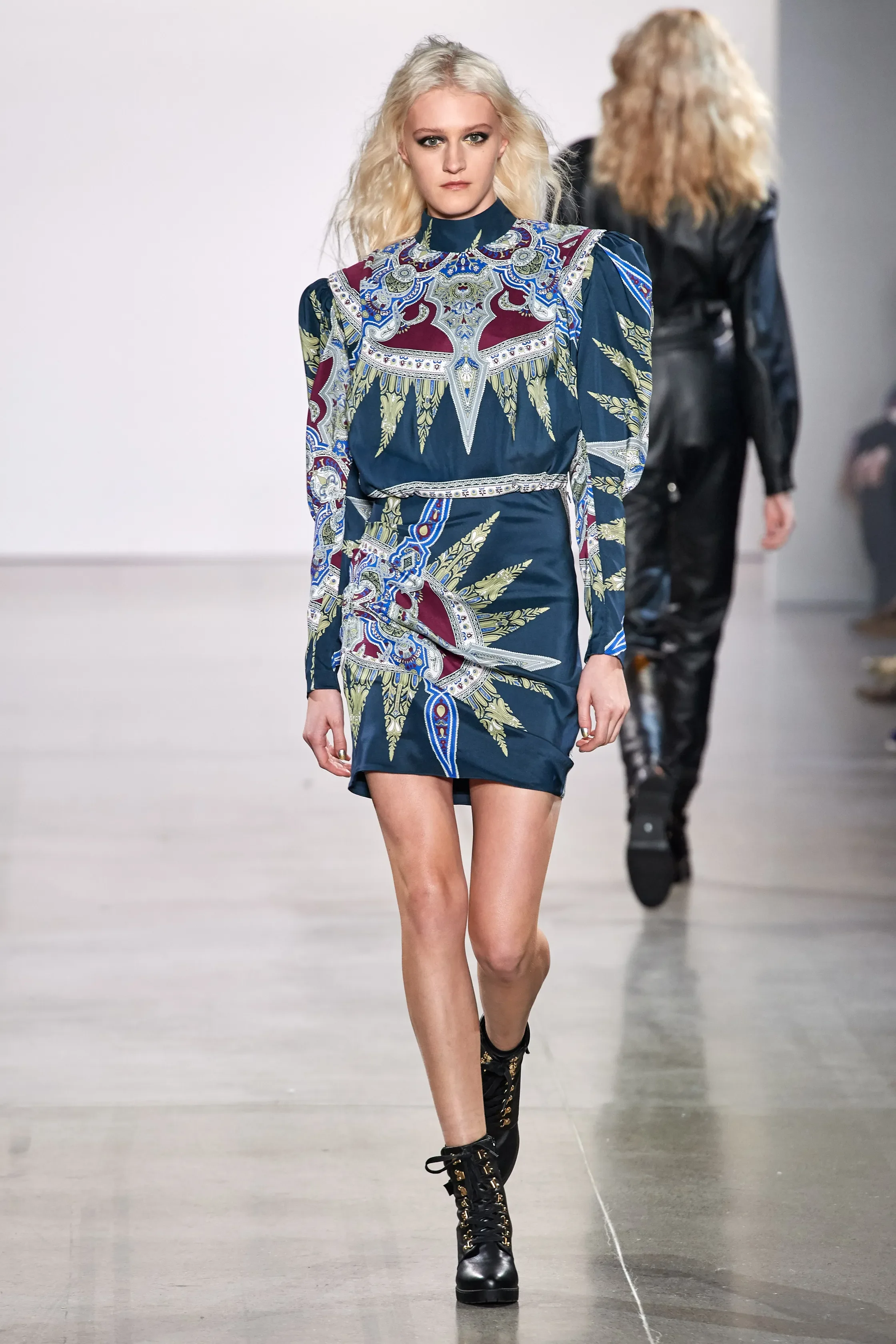 A female model with blonde hair walking on a runway, wearing a colorful patterned dress and black lace-up ankle boots.