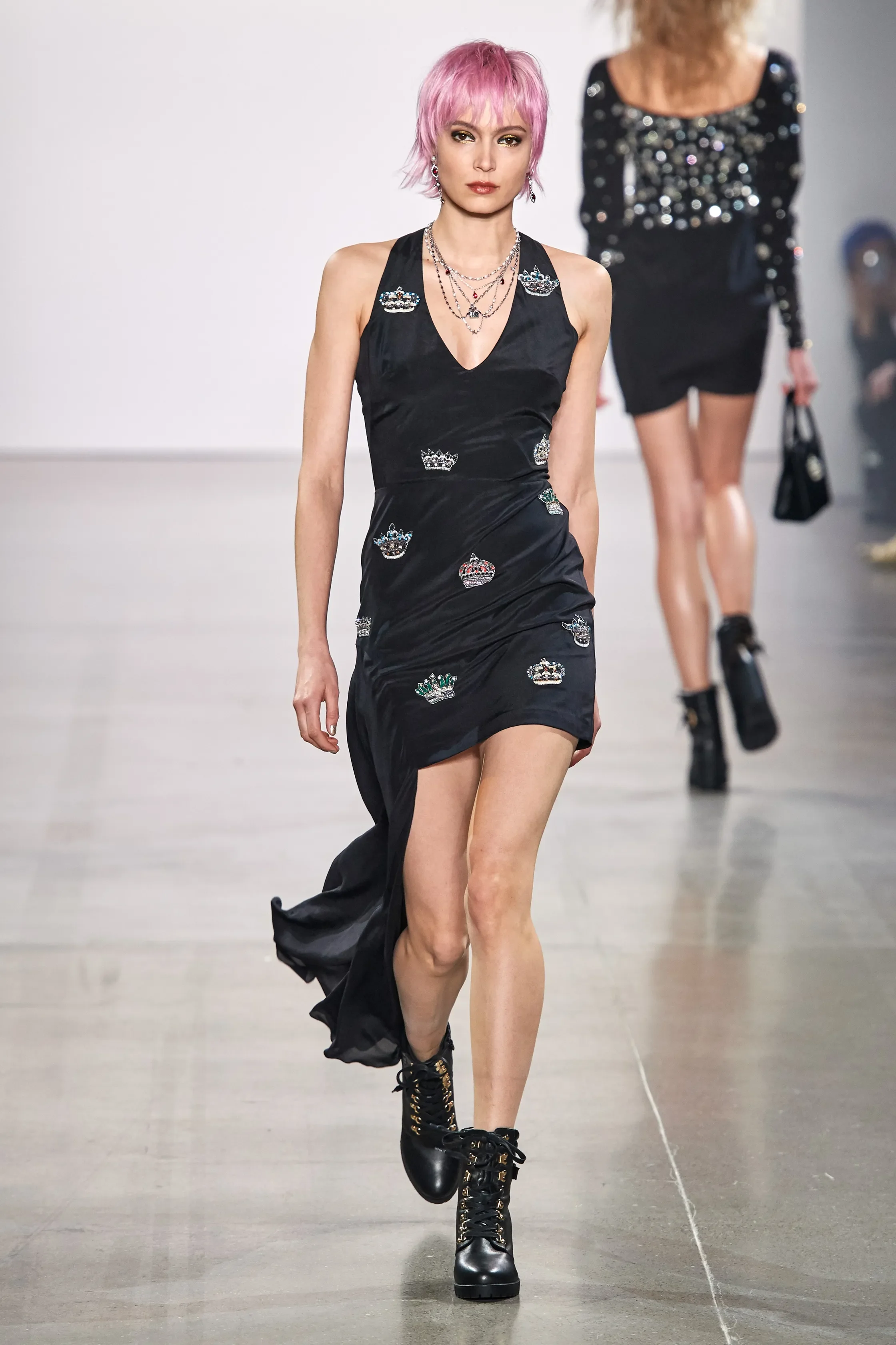 A fashion model with pink hair walks down the runway wearing a black dress adorned with colorful crown embellishments, paired with black combat boots.