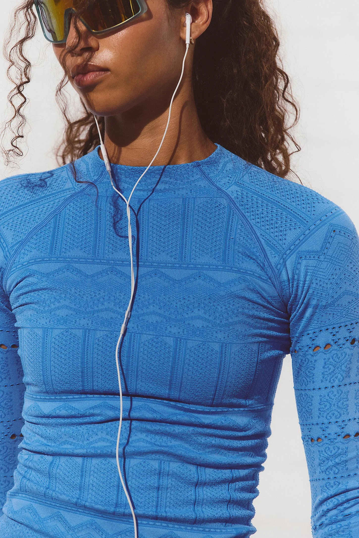 Close-up of a woman wearing black sports sunglasses, a blue athletic top with intricate patterns, and white earphones, against a plain background.