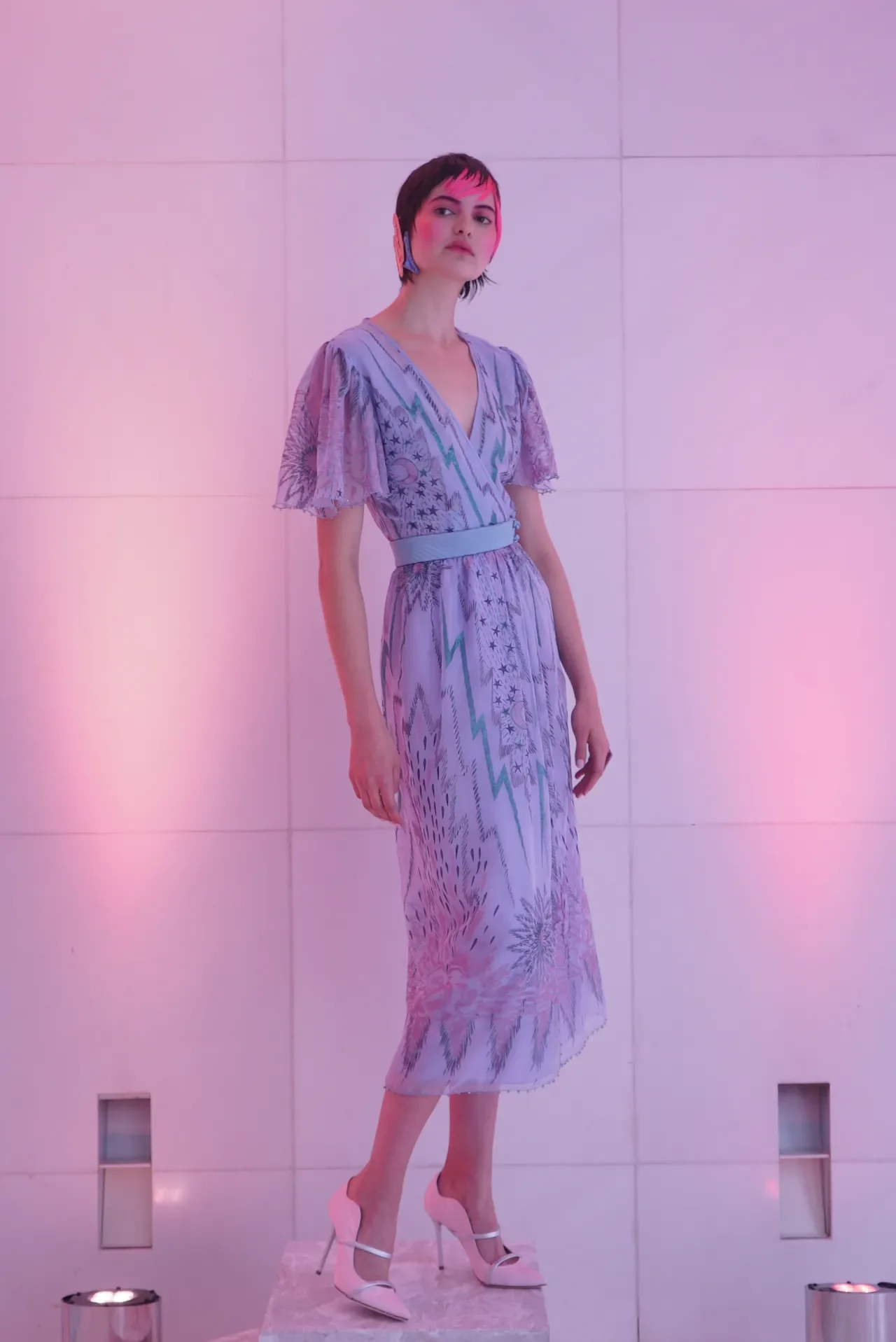 A woman with short dark hair and pink makeup, standing in front of a pink-lit wall, wearing a light purple patterned dress and white high heels.