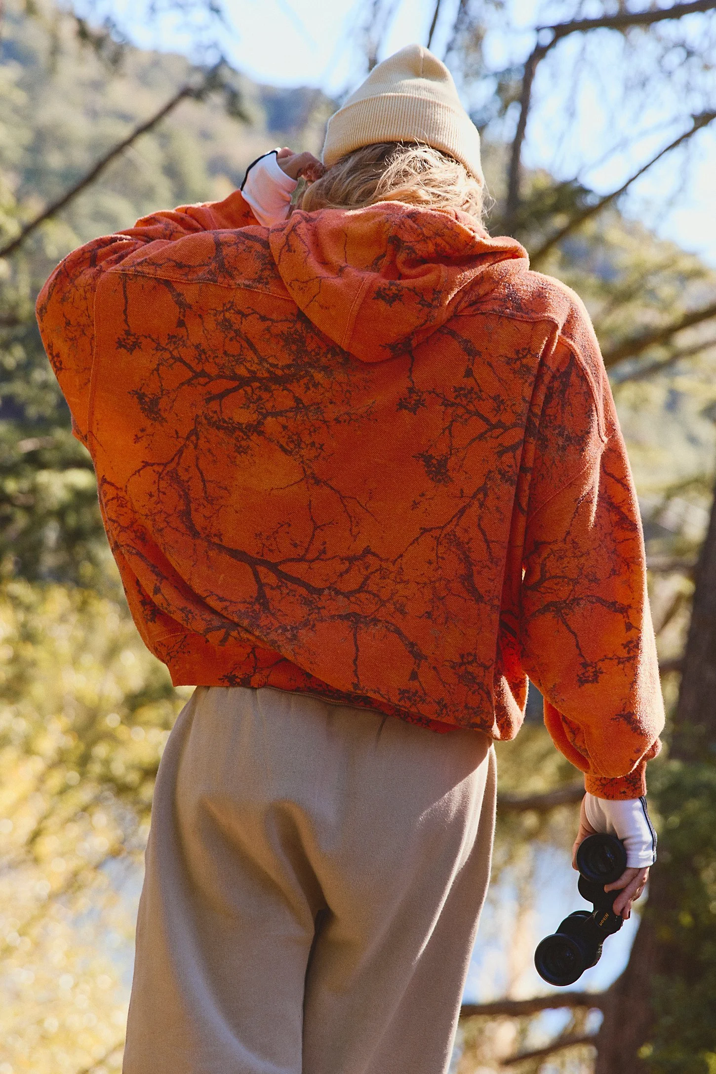 Person wearing an orange tie-dye hoodie, holding binoculars, outdoors in a wooded area with trees and blue sky in the background.
