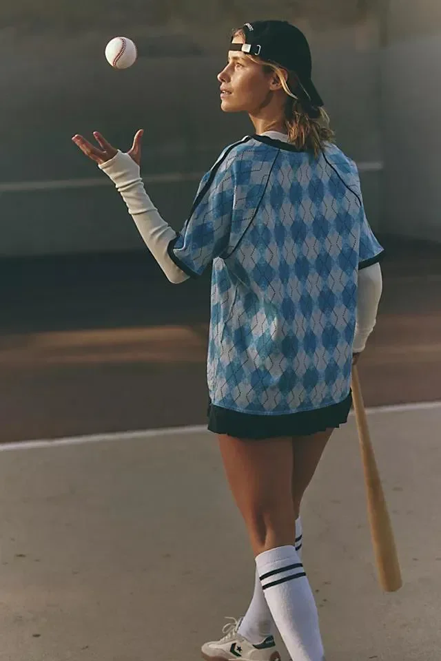 A young woman with light brown hair wearing a backwards black baseball cap, a blue argyle baseball jersey, white long-sleeve shirt underneath, black shorts, white knee-high socks with black stripes, and white sneakers, standing on a baseball field to