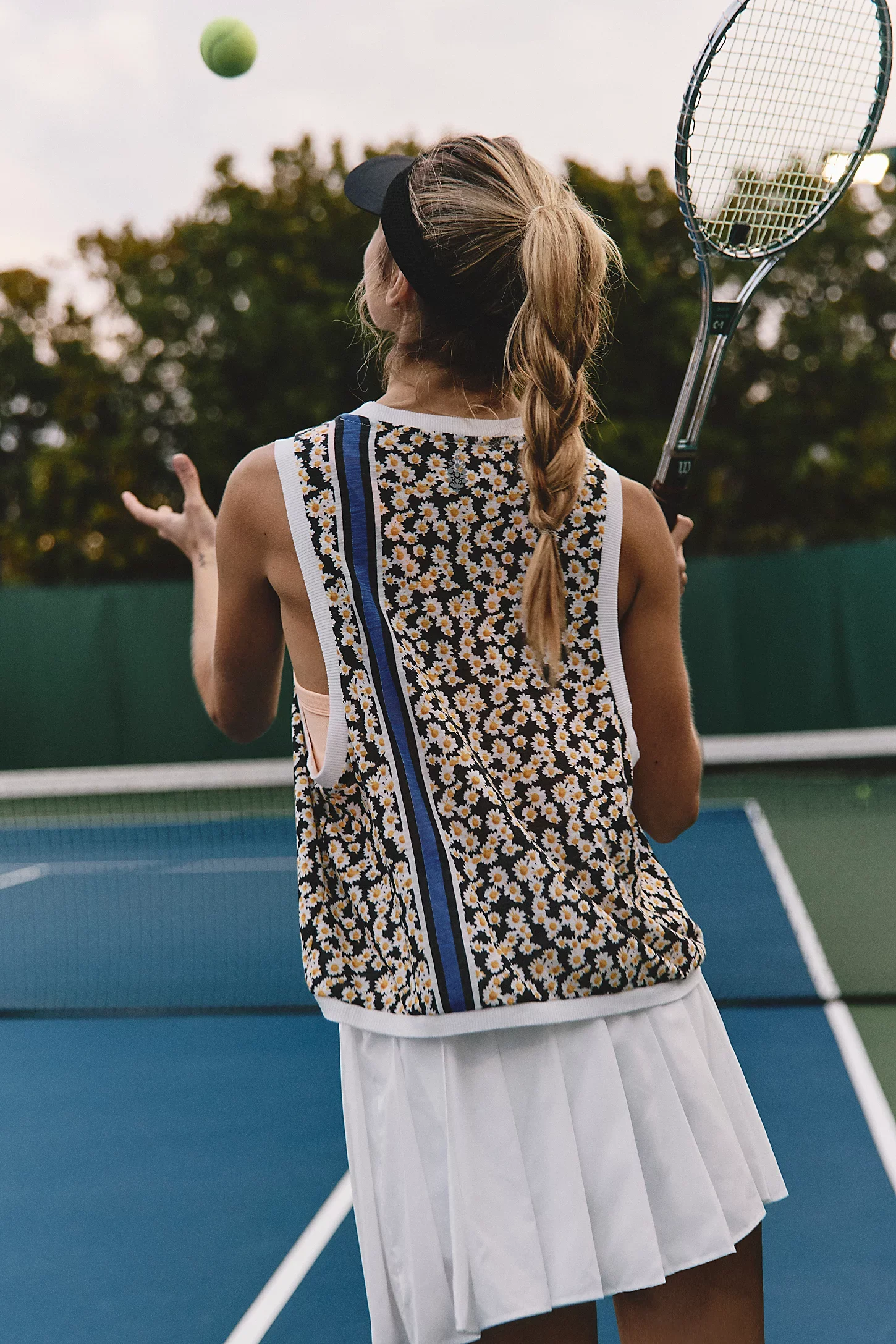 A young girl playing tennis on an outdoor court, holding a tennis racket and preparing to hit a tennis ball, with trees in the background.