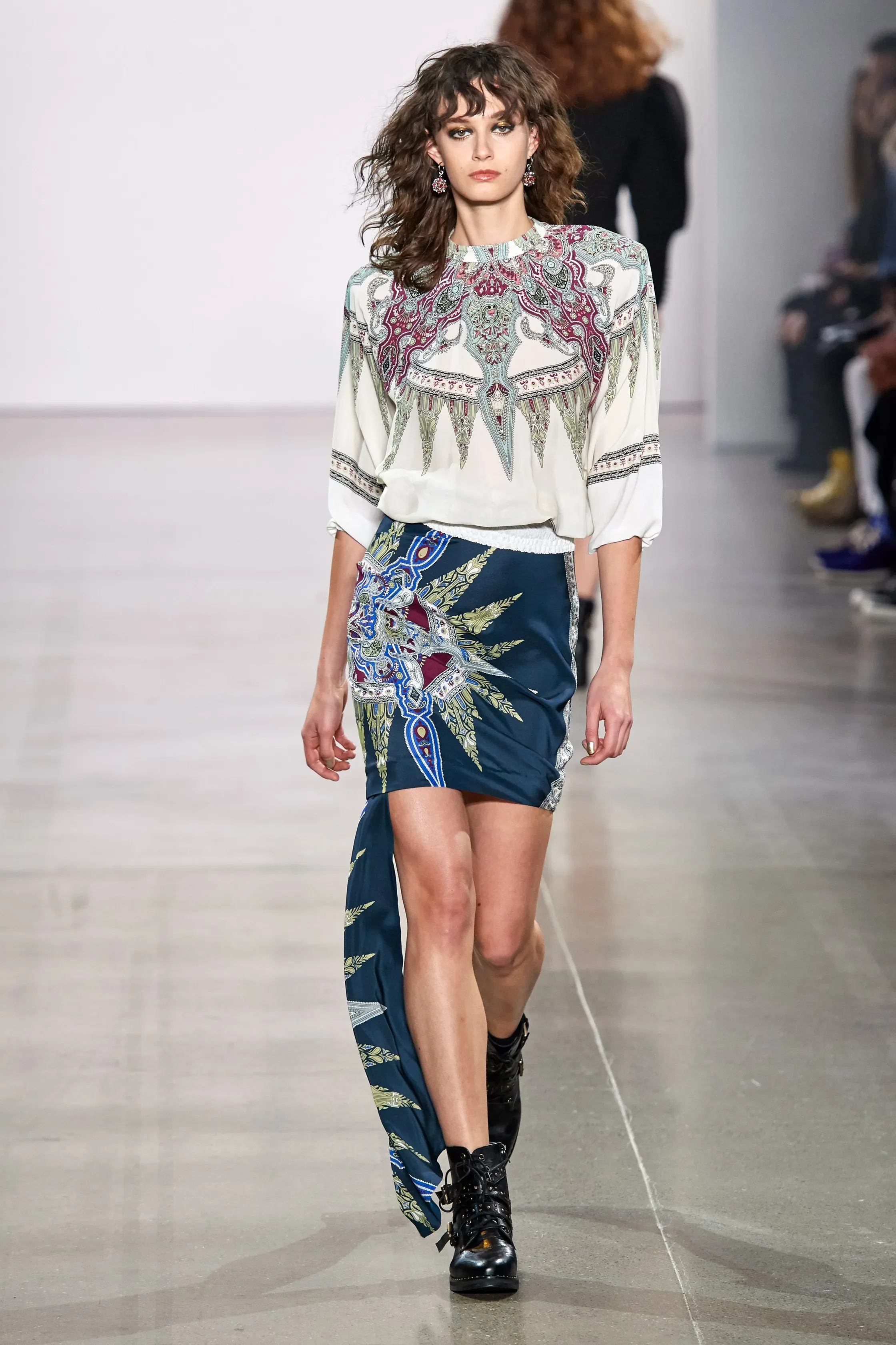 A female model walking on a runway during a fashion show, wearing a printed blouse and a matching skirt with a slit, paired with black combat boots.