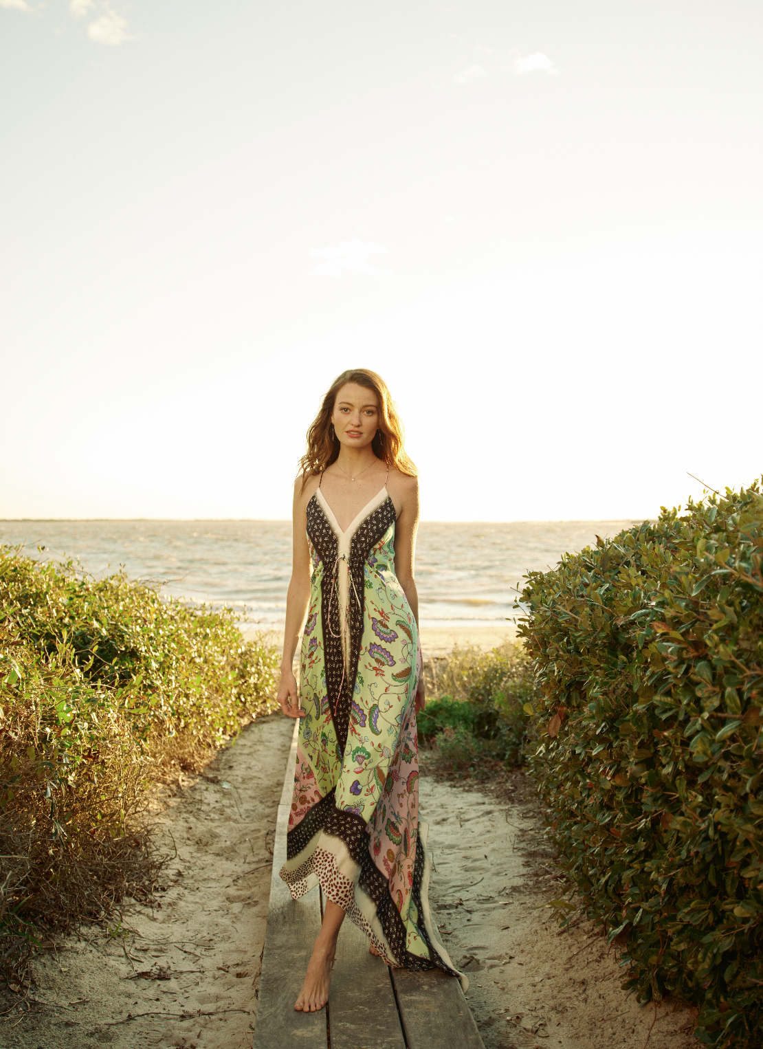 A woman in a colorful dress walking on a wooden path through bushes towards the beach during sunset.