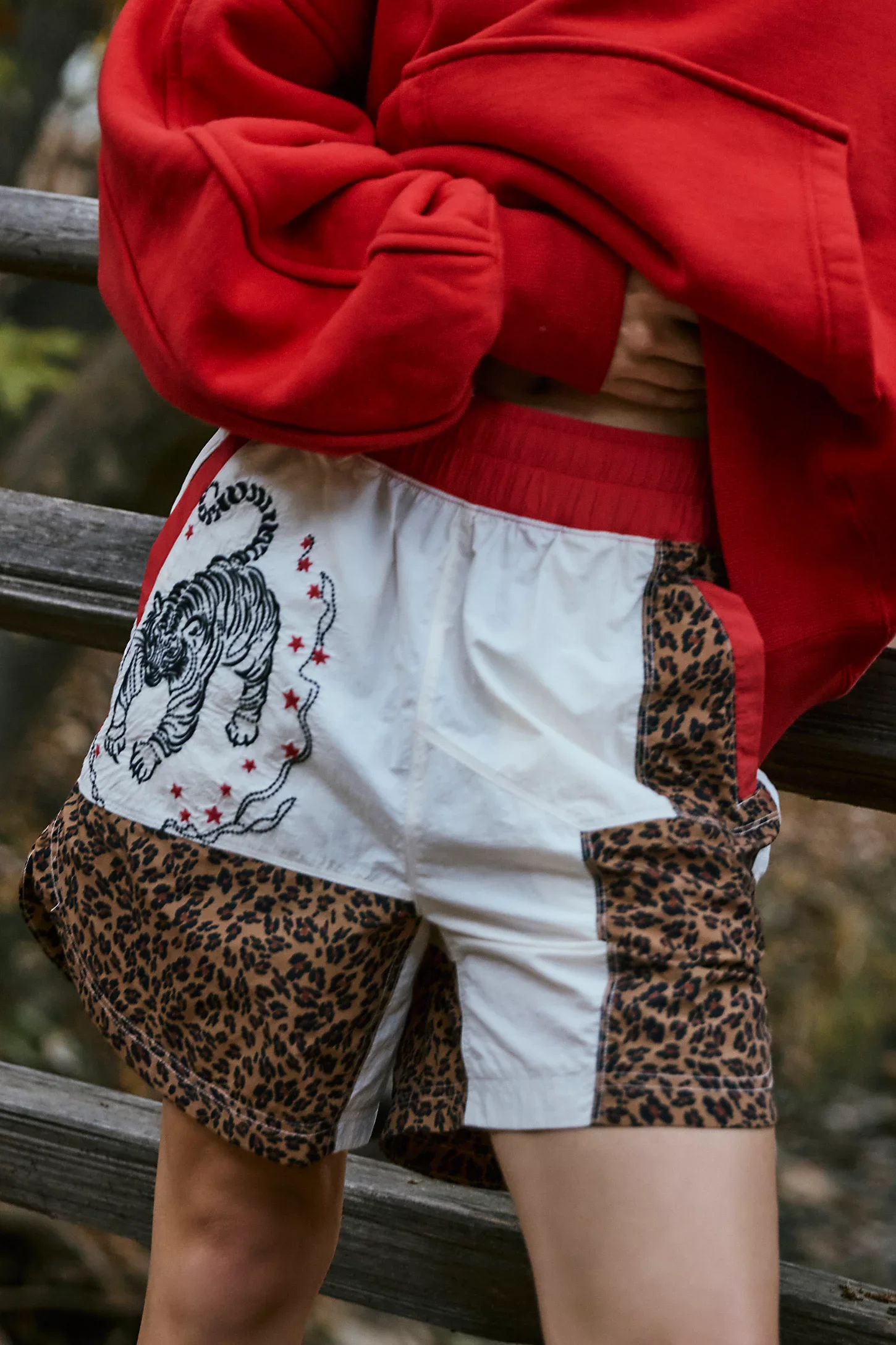 Person wearing a red hoodie and shorts with leopard print and tiger design, standing outdoors near a wooden railing.