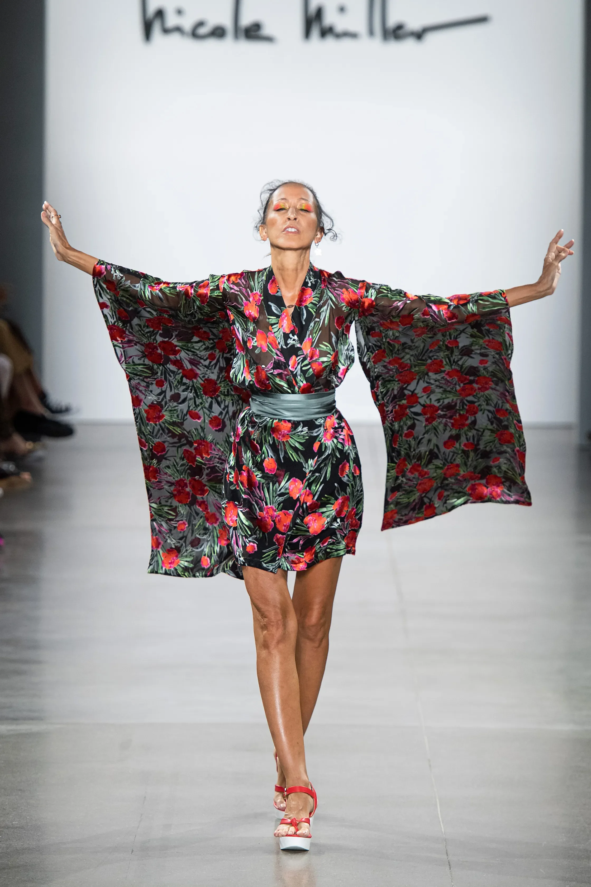 Model walking on runway in floral kimono-style dress with arms outstretched, audience on the side, designer's name 'Nicole Miller' displayed in the background.