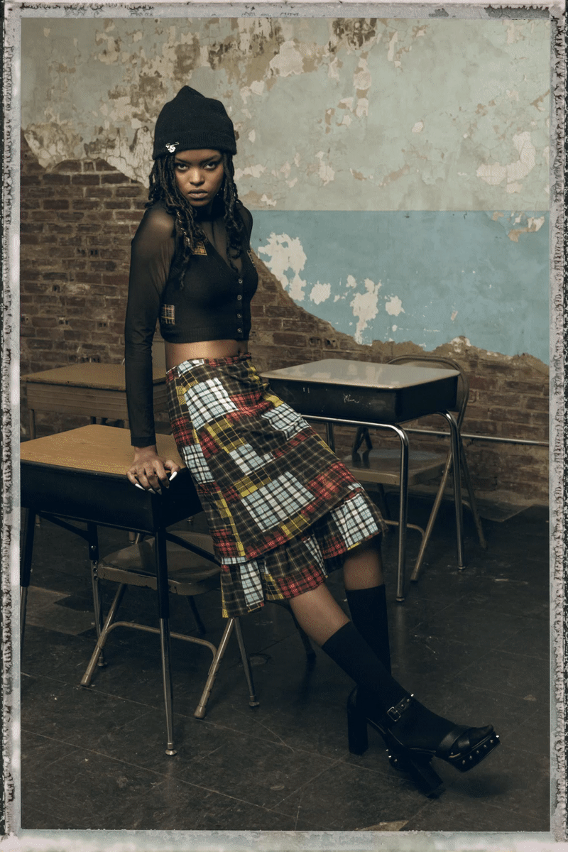 Young woman sitting on a table in a vintage room with peeling paint and brick wall, wearing a black beanie, black sheer top, plaid skirt, and platform heels.