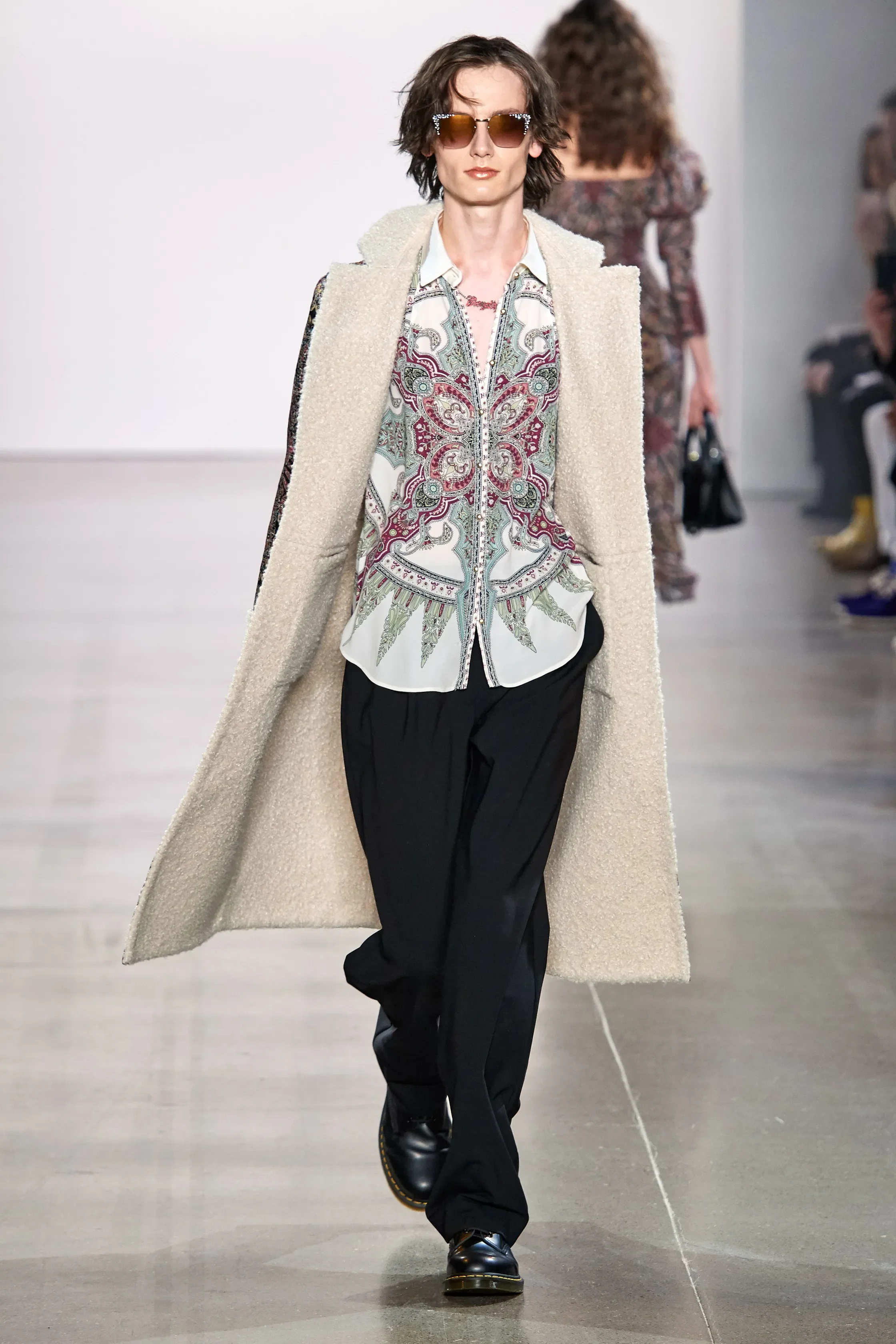 Model walking down runway wearing a long beige coat, embroidered blouse, black pants, sunglasses, and black shoes at fashion show.
