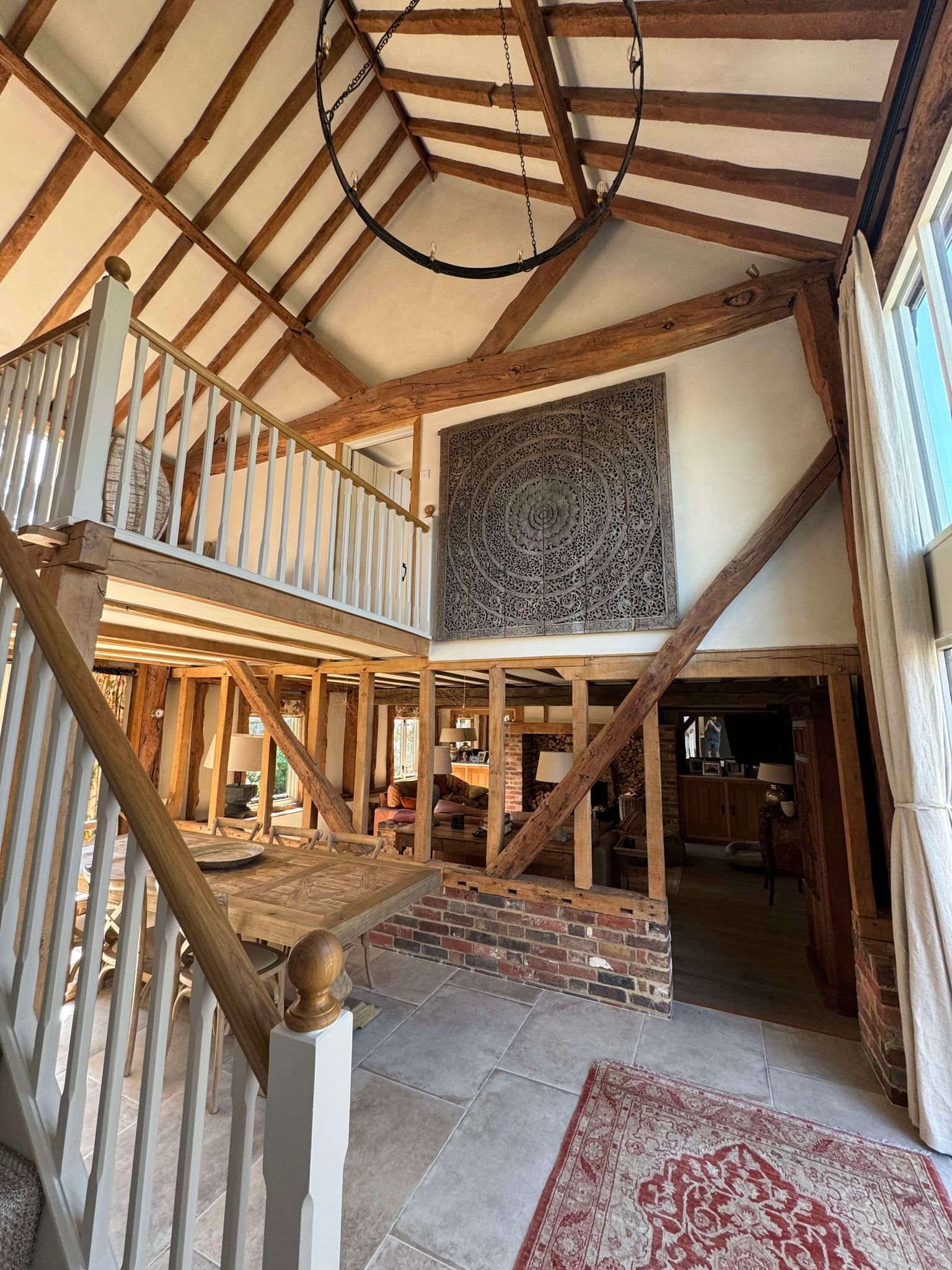 Interior view of a rustic home with exposed wooden beams, a high ceiling, a hanging circular chandelier, stairs with a white railing, and a large decorative wall hanging. The space includes a dining area with a wooden table, a cozy sitting area with chairs and a fireplace, and large windows with curtains.
