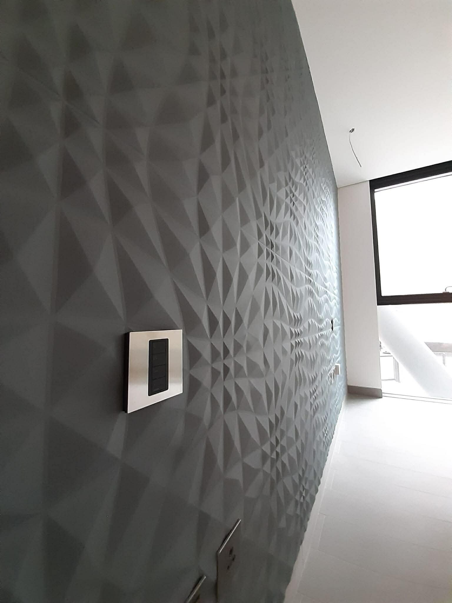 Interior wall with a 3D textured geometric pattern in shades of gray, with electrical outlets and a large window providing natural light.
