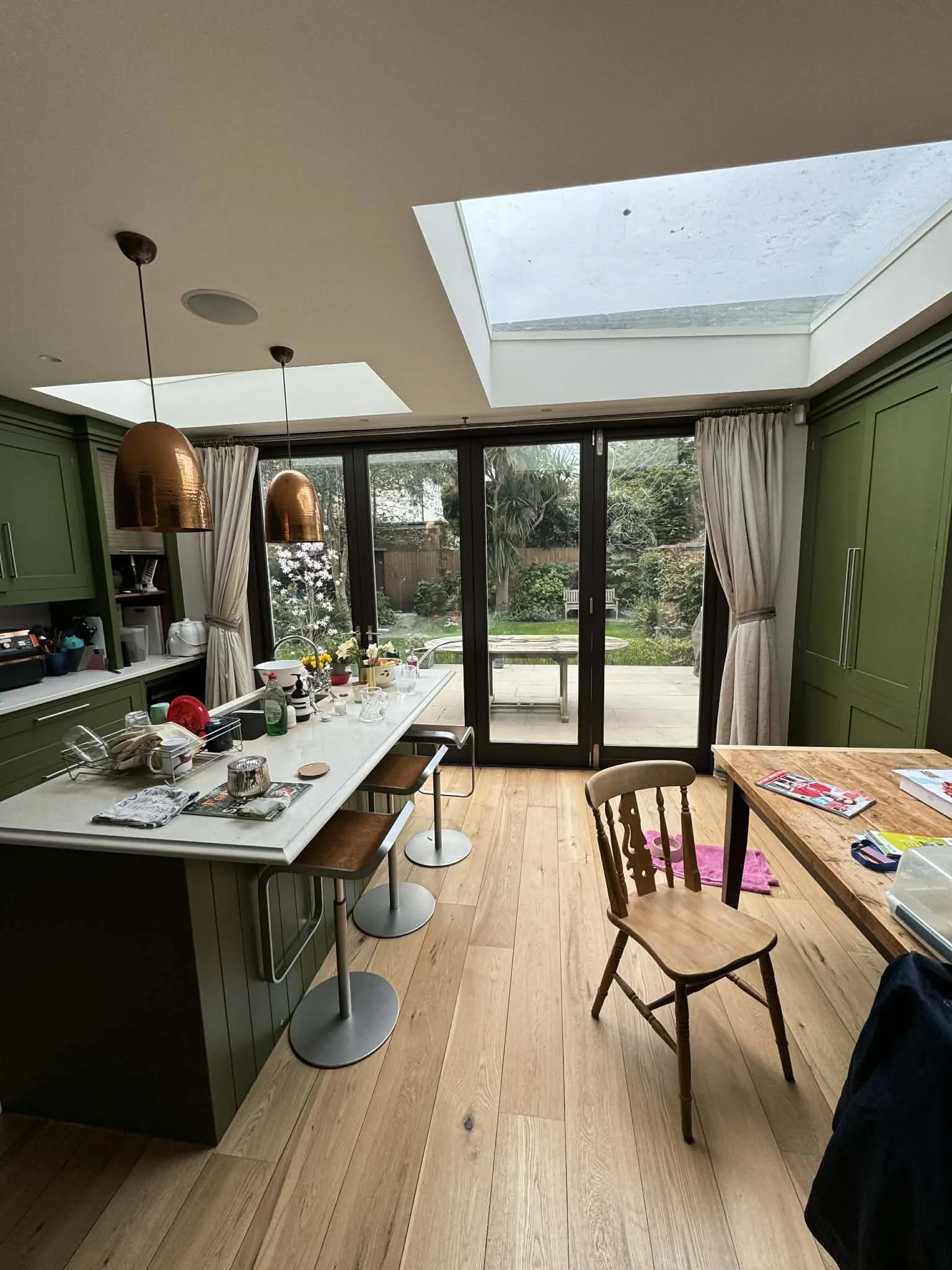 A kitchen with green cabinets, a white island with bar stools, wooden dining table and chair, sliding glass doors with curtains, and large skylights in the ceiling, overlooking a backyard garden.
