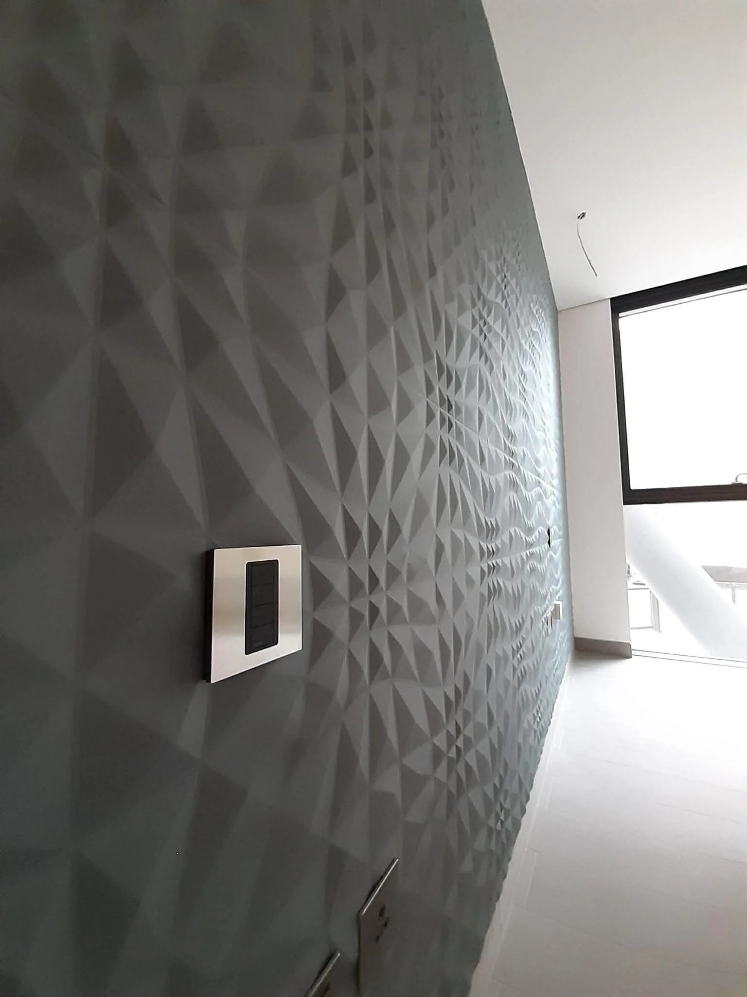 Close-up of a modern textured wall with a geometric pattern in a bright room, including a control panel and electrical outlets.