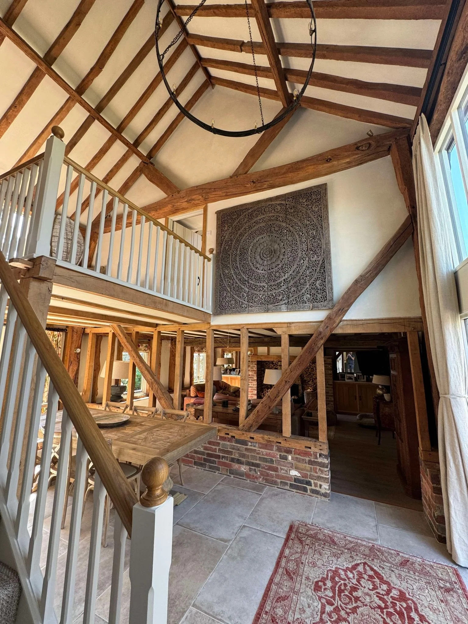 Interior view of a rustic, two-story home with exposed wooden beams, a loft area with a white railing, a large framed decorative wall hanging, and a glimpse into the living room with furniture and a fireplace.