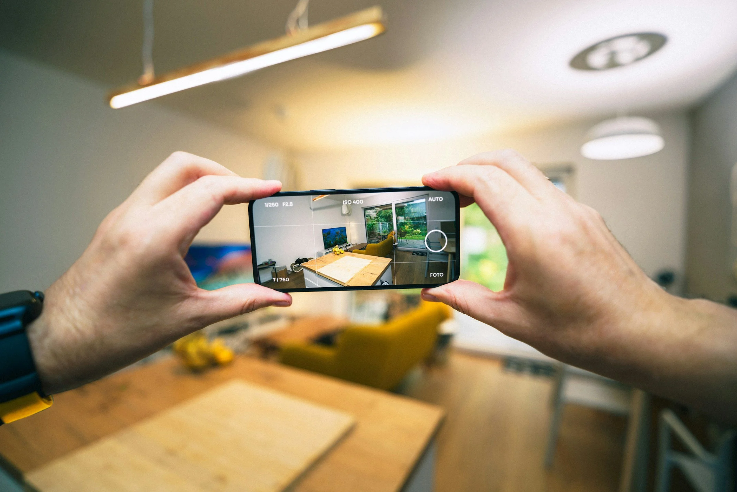 Person taking a photo of a kitchen and dining area with a smartphone camera, showing a wooden dining table, yellow chairs, a TV on the wall, and large windows revealing a garden outside. On TOGAR & Co.'s Real Estate Website.