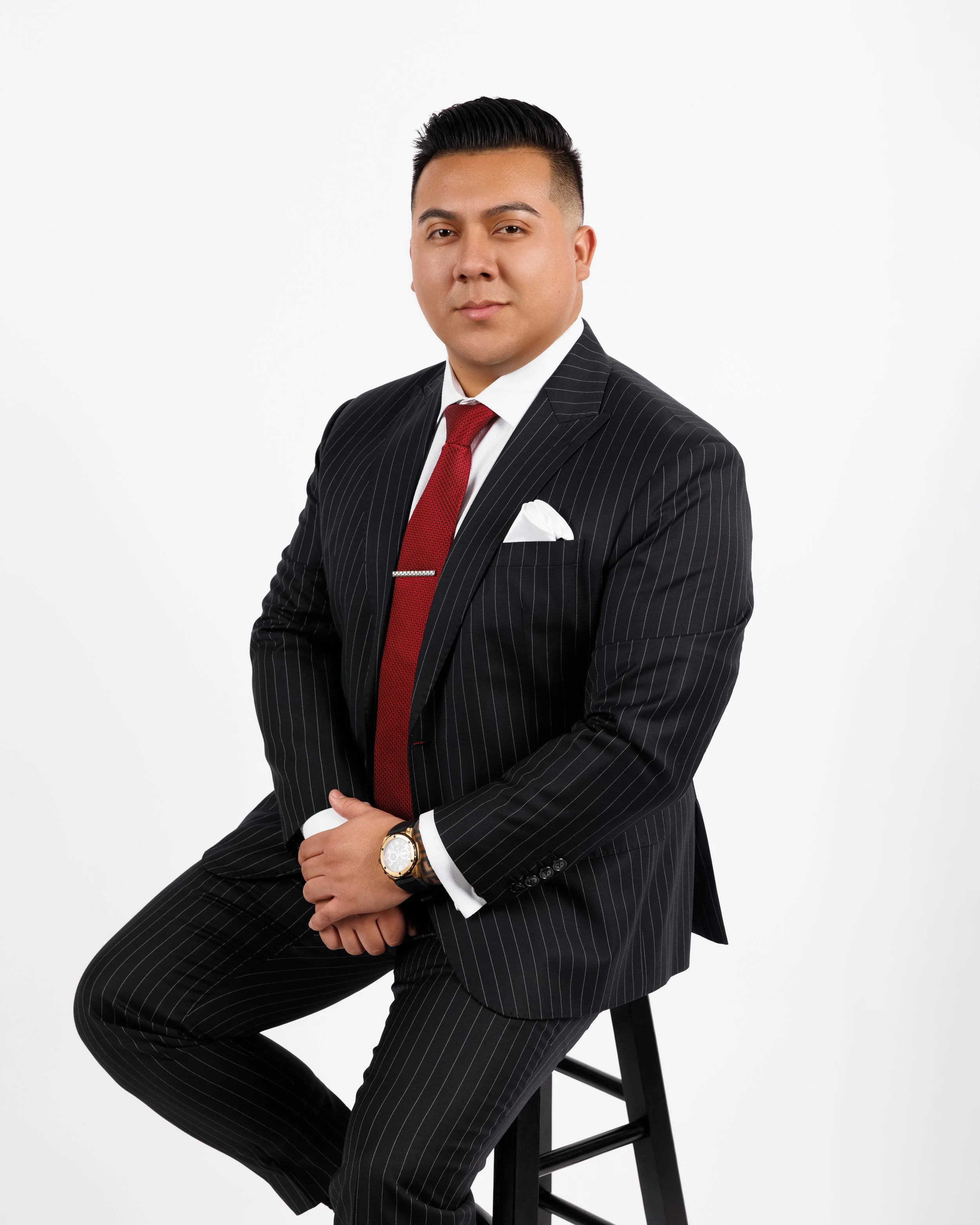 TOGAR & Co.'s Founder, Tony Garcia. A real estate agent in a black pinstripe suit with a white shirt and red tie, sitting on a stool against a plain white background.