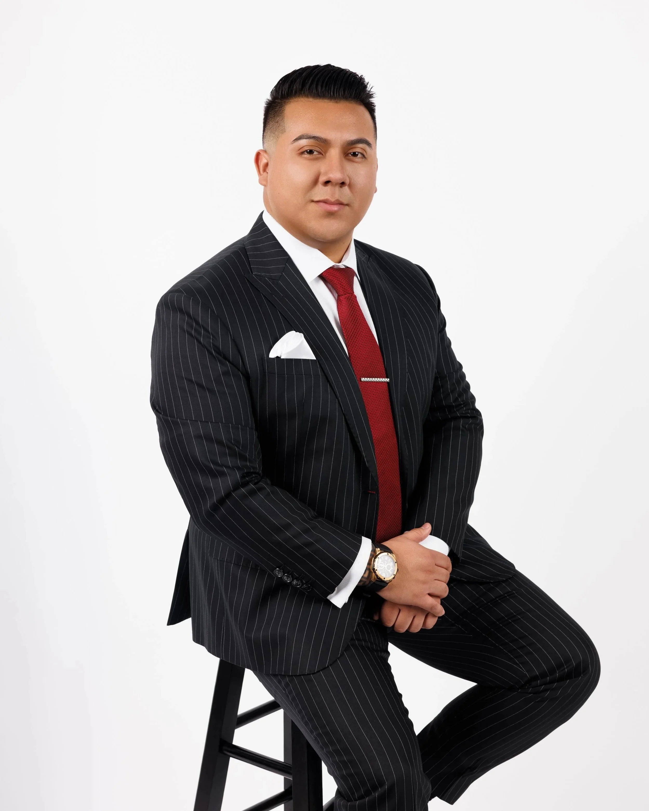 Tony Garcia Chicago Realtor Portrait. A man dressed in a black pinstripe suit with a white shirt and red tie sitting on a black stool against a white background. TOGAR & Co. Chicagoland Real Estate
