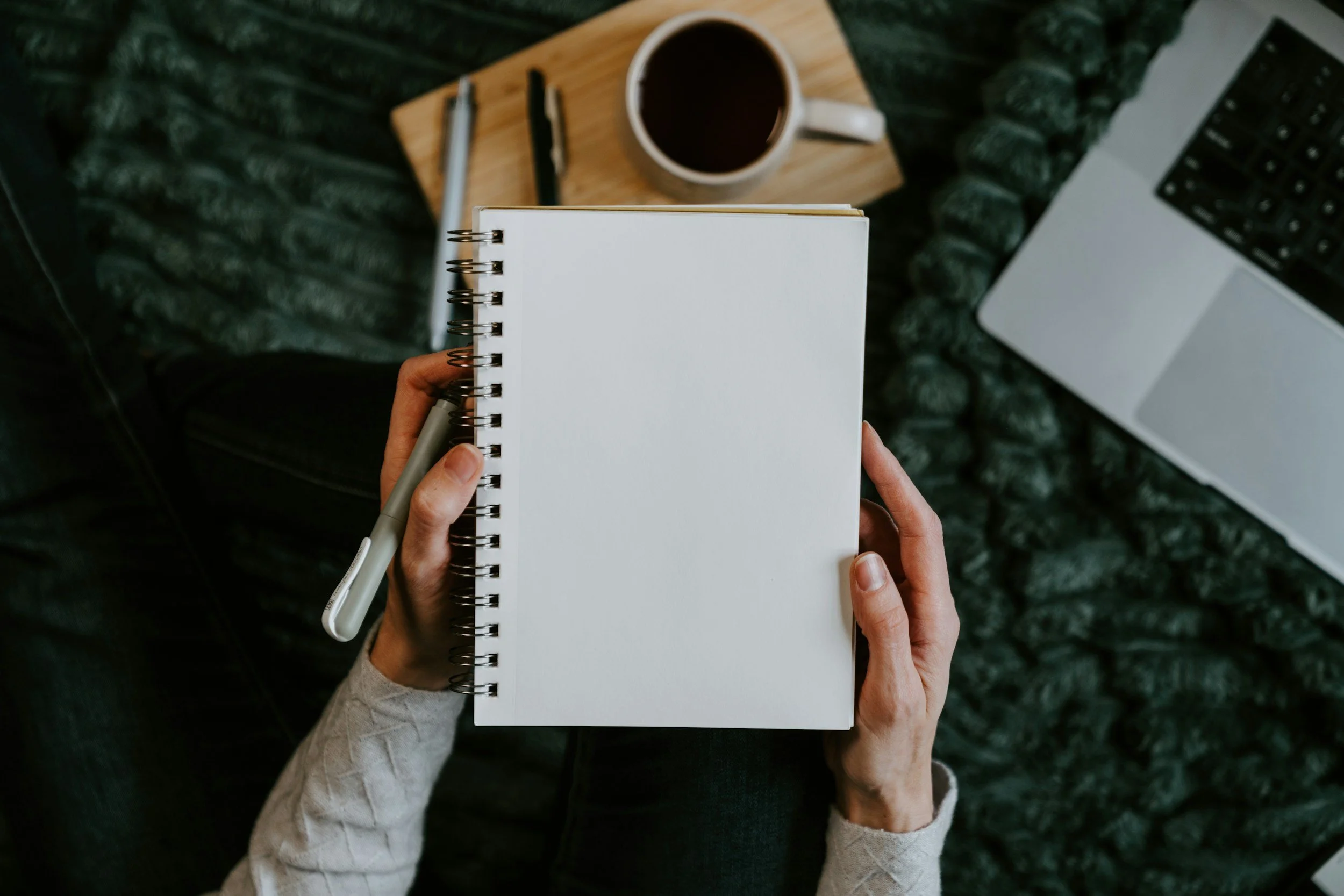 A person holding a blank spiral notebook and pen, sitting at a desk with a coffee mug, pens, a small wooden board, a laptop, and a mouse.