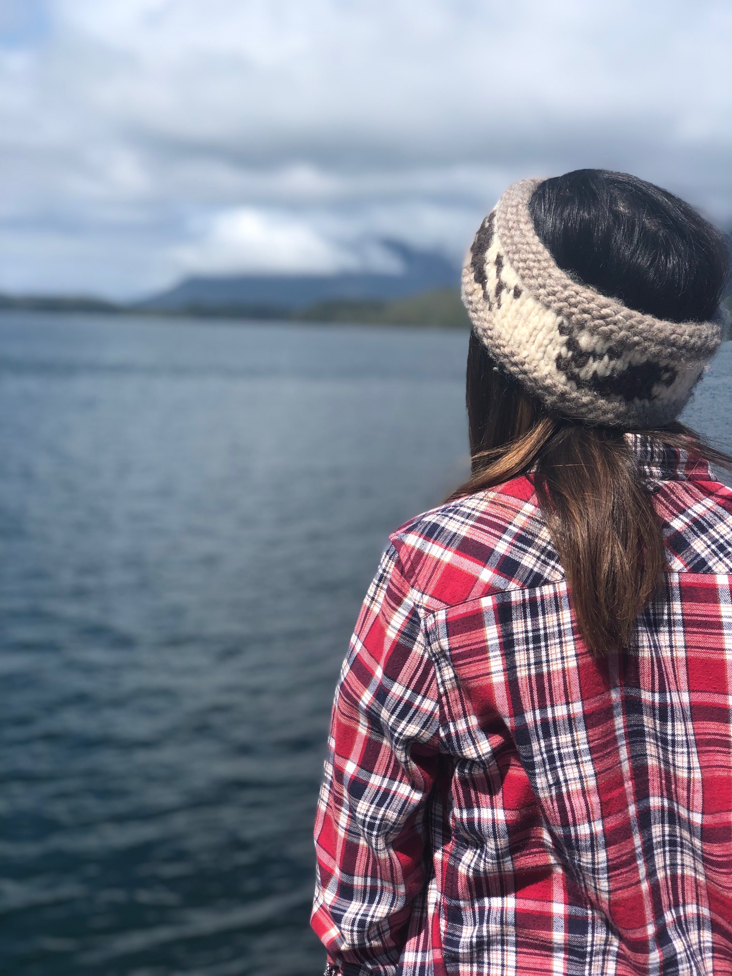 A woman wearing a wool headband and a red plaid jacket standing outdoors by a body of water, with mountains and cloudy sky in the background.