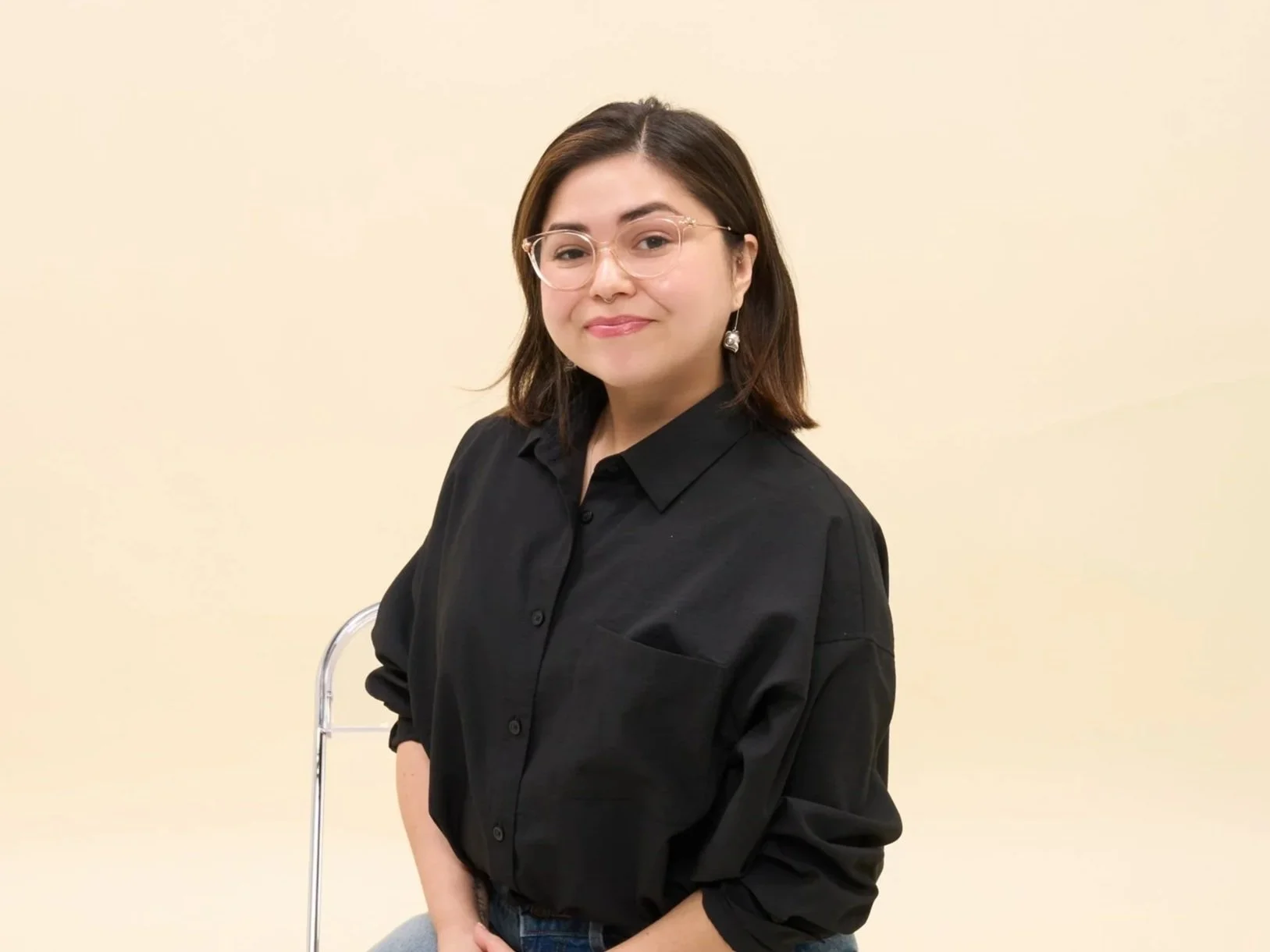 A woman with shoulder-length dark hair, wearing glasses and earrings, is smiling and standing against a plain beige wall, dressed in a black button-up shirt.