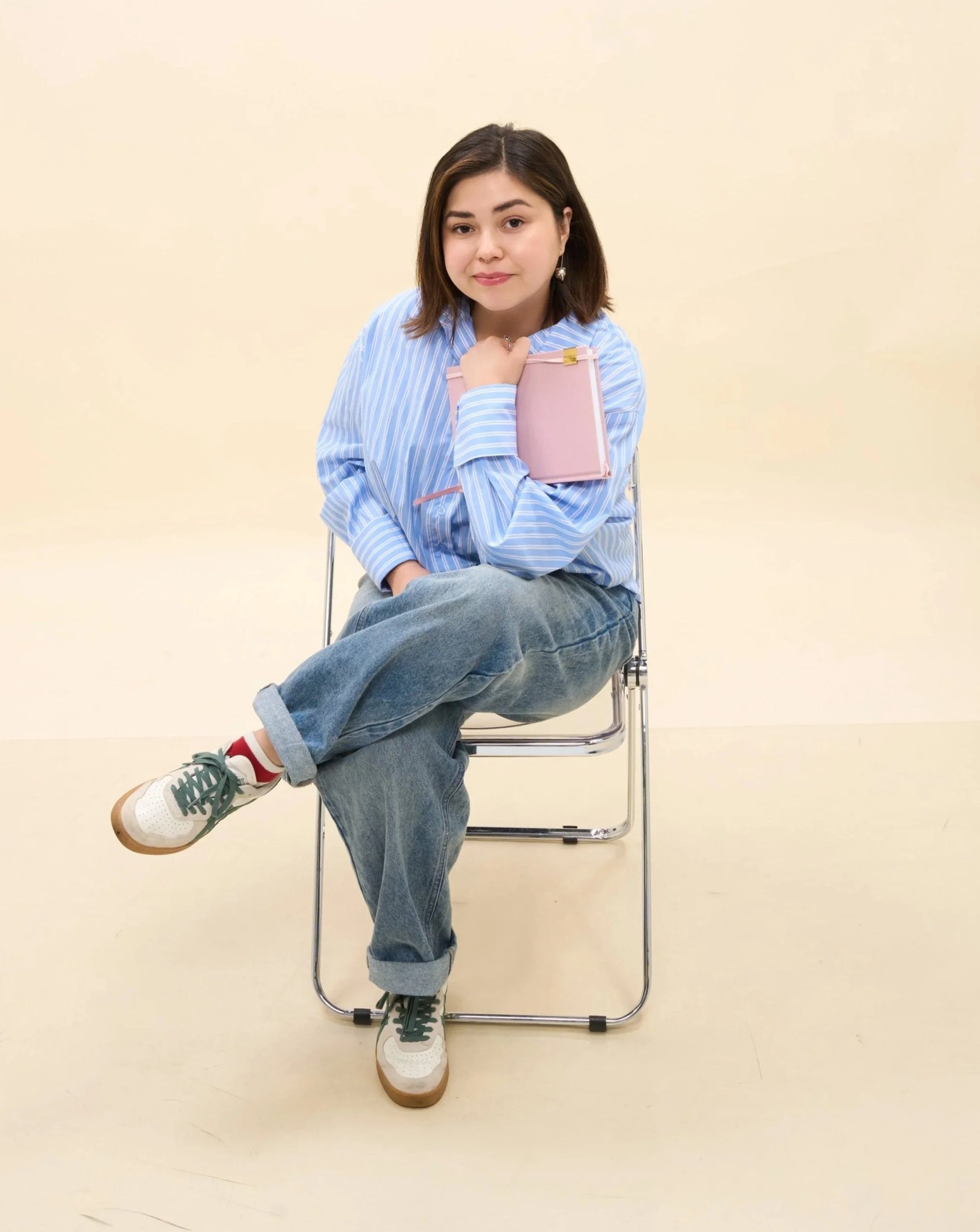 A woman sitting on a metal chair with crossed legs, wearing a blue and white striped shirt, jeans, and sneakers, holding a pink notebook on her chest, with a neutral expression and short brown hair.