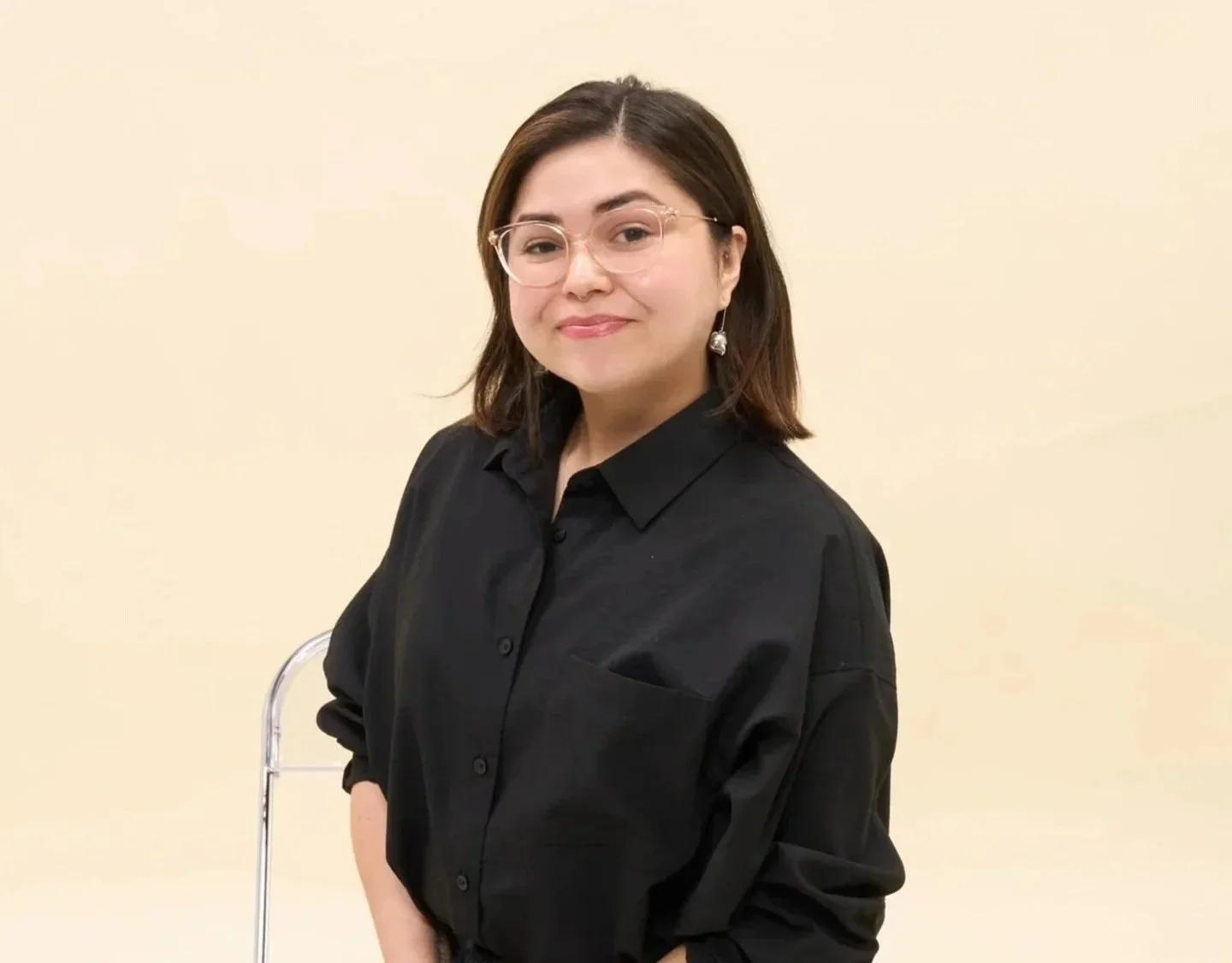 A woman with glasses and earrings, wearing a black button-up shirt, standing against a light beige background, smiling at the camera.