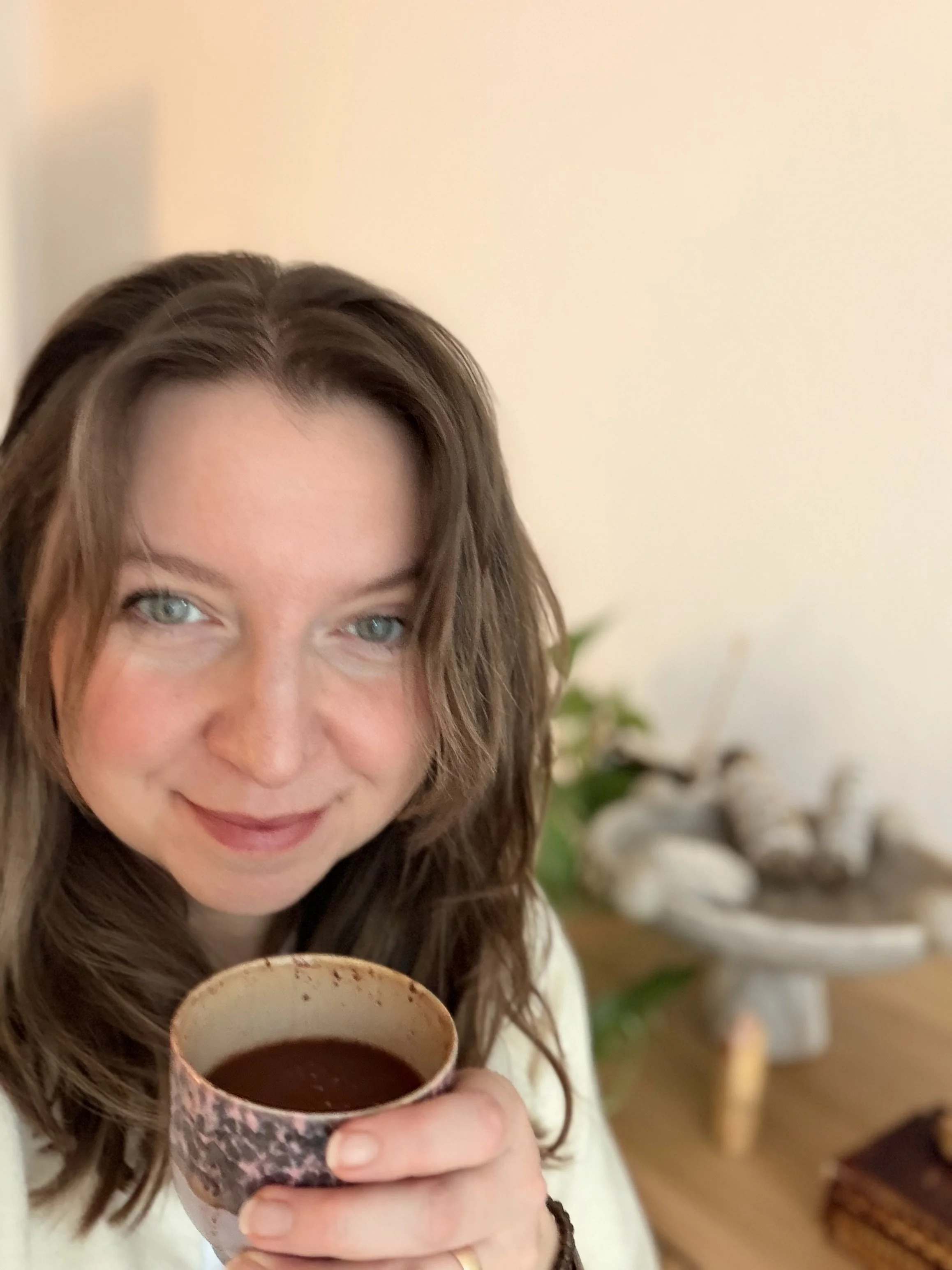 A woman with brown hair holds a cup of cacao and smiles, in a room with a plant, decorative stones, and a table in the background.