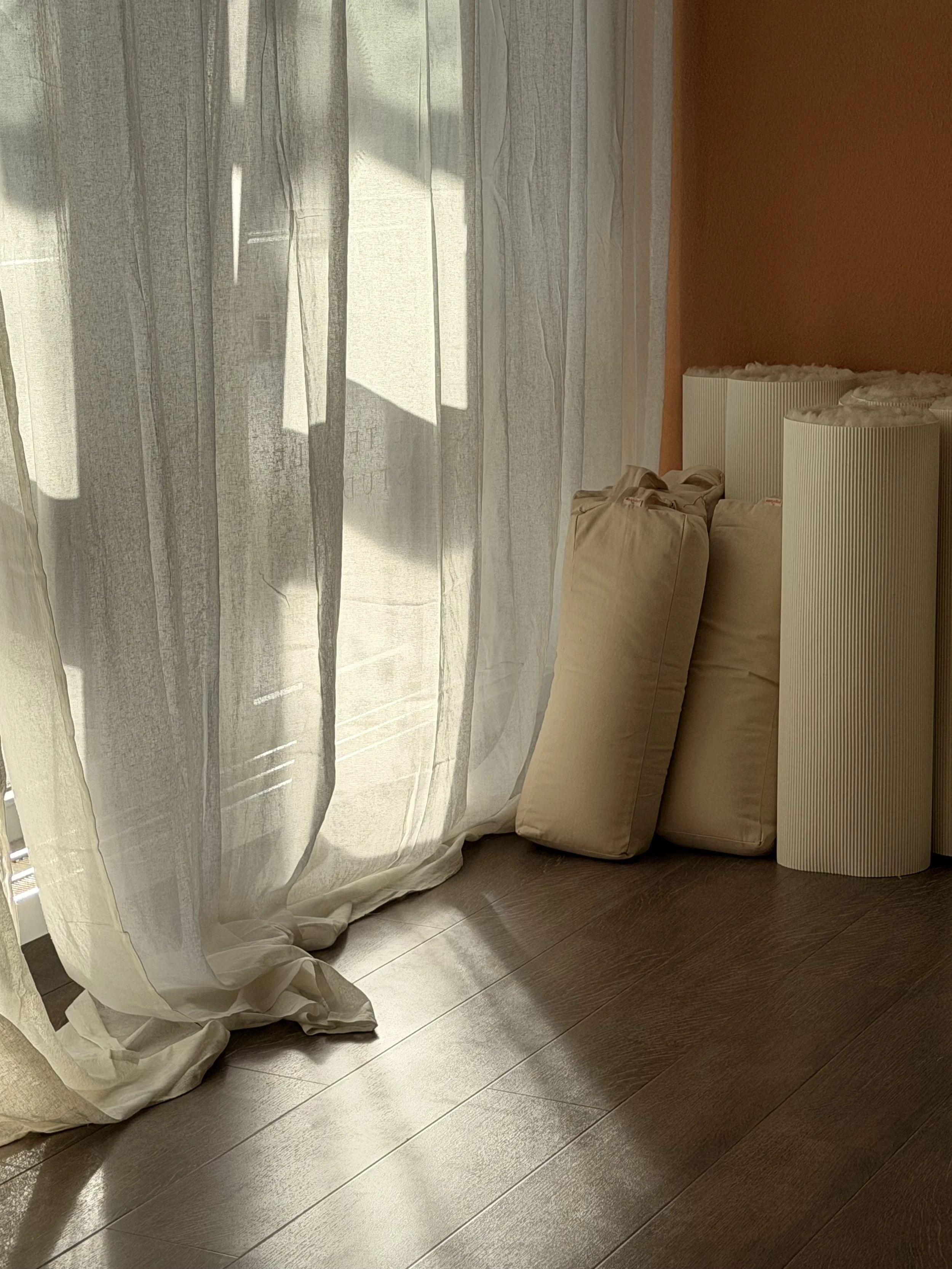 Blick auf einen sonnendurchfluteten Raum mit weißen Vorhängen, beige Bolstern und Schaffellmatten auf Holzboden.