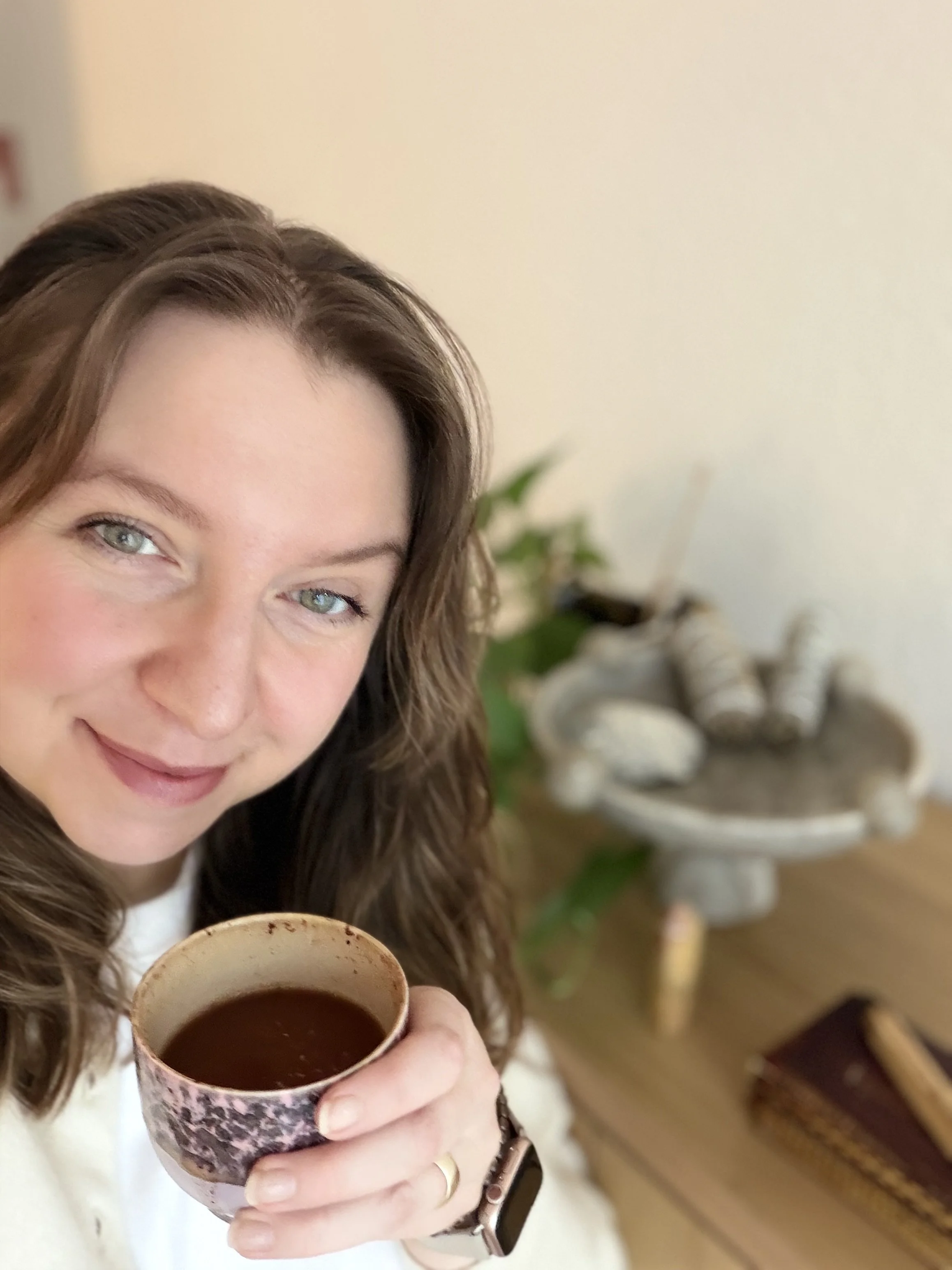 A woman with long brown hair and blue eyes holding a cup of ceremonial cacao inside a room with a blurred background that includes a plant, a decorative fountain, and a wooden table.