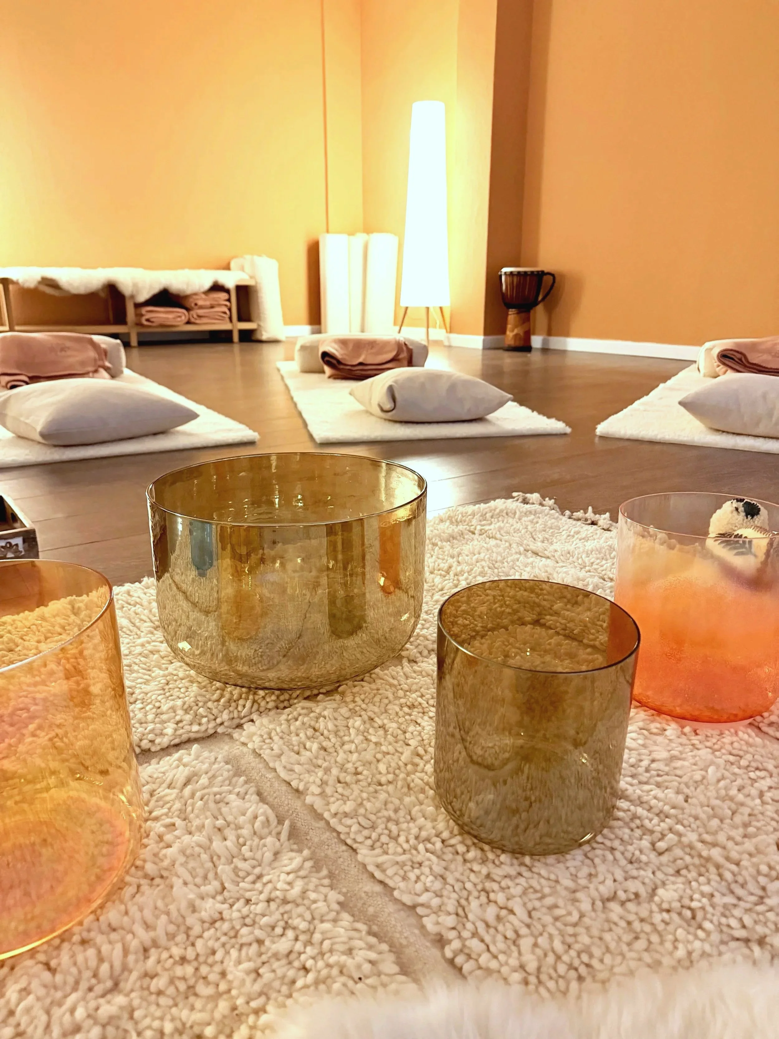 A cozy room with mats, pillows, and blankets arranged on the floor, illuminated by a tall floor lamp, with a drum in the background and crystal singing bowls on a beni ourain in the foreground.