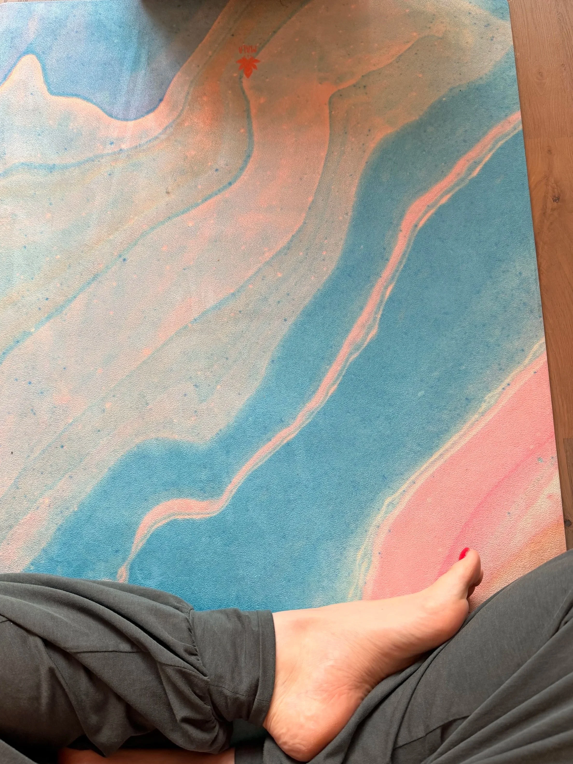 Eine Person sitzt auf einer blau-rosanen Yogamatte, trägt eine grau-grüne weiche Hose und hat rote Fußnägel. Die Person scheint eine entspannte Haltung einzunehmen.