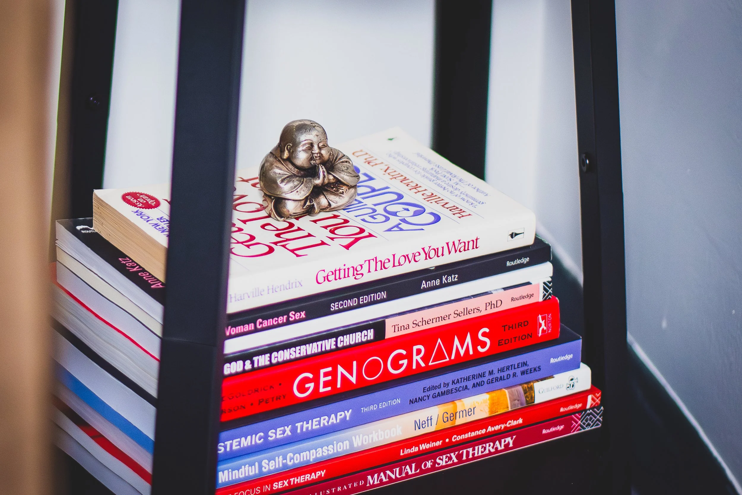 Stack of books on a black shelf, including titles on love, sex, reproductive health, and therapy, with a small metallic figurine of a person sitting with hands clasped in front.