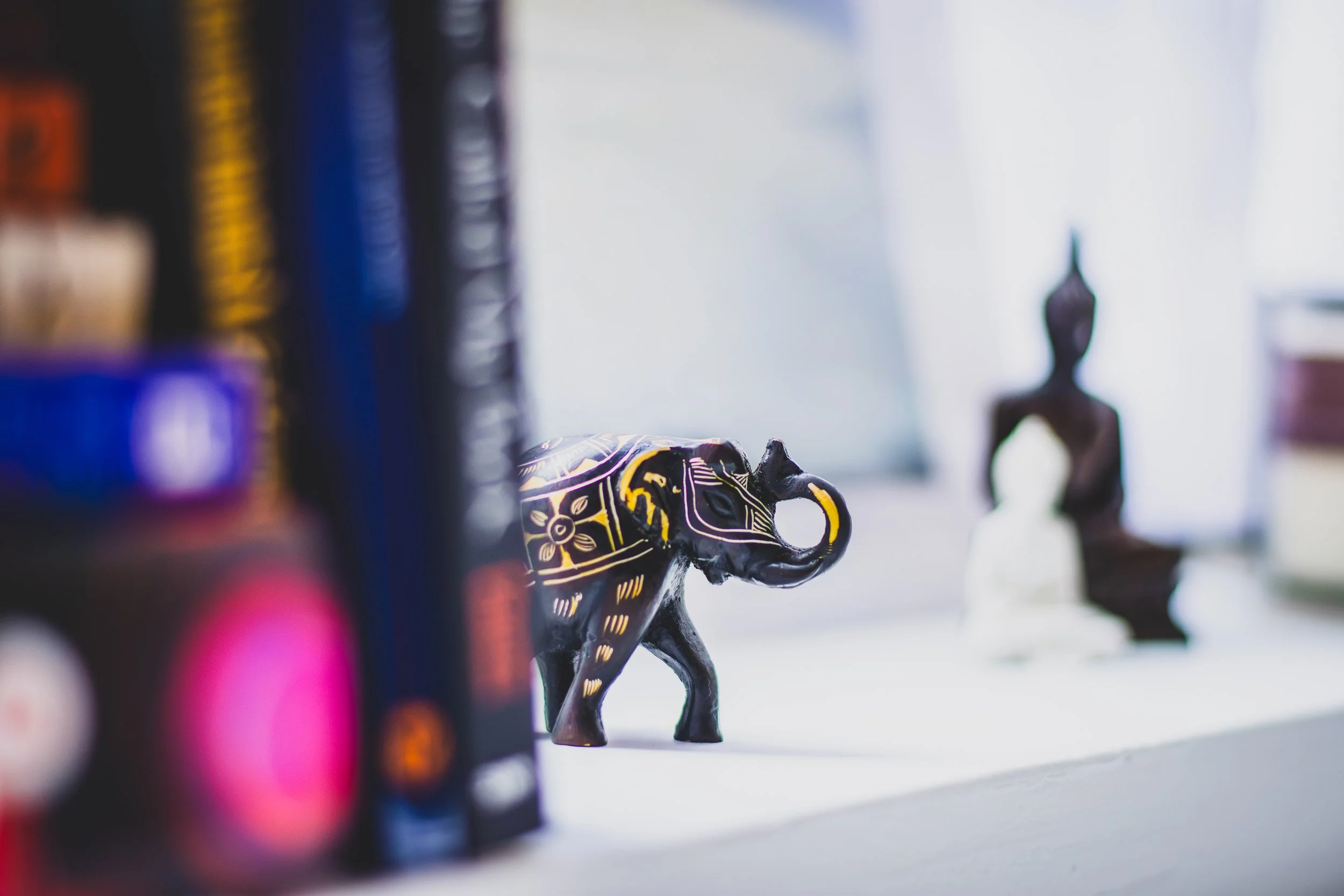 Decorative black elephant figurine with yellow and purple patterns on a white surface, with blurred book spines on the left and two small chess piece statues in the background on the right.