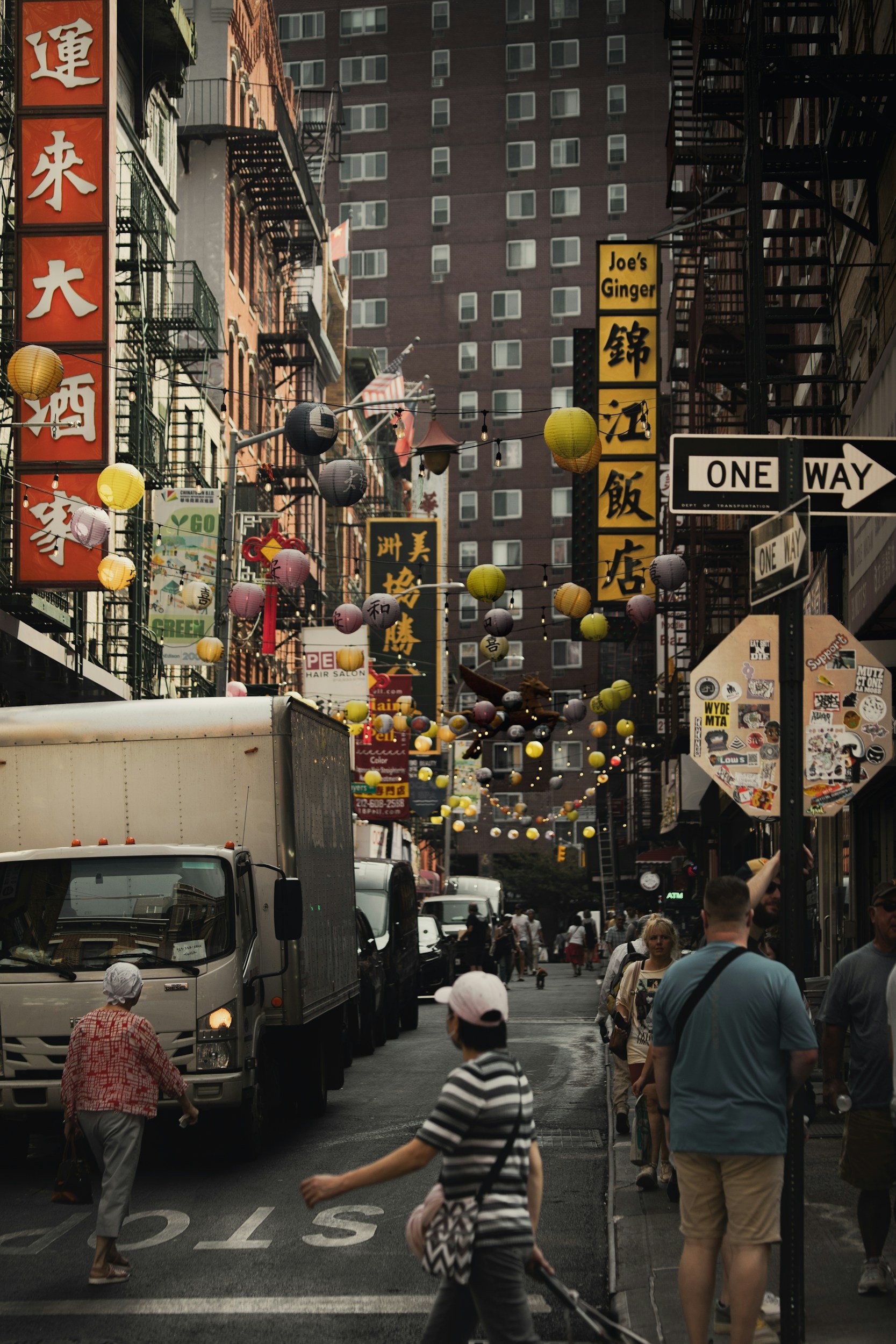 City street scene with people walking and trucks parked along the side. Colorful paper lanterns hang across the street, with Chinese and English signs on buildings. Tall buildings tower in the background.