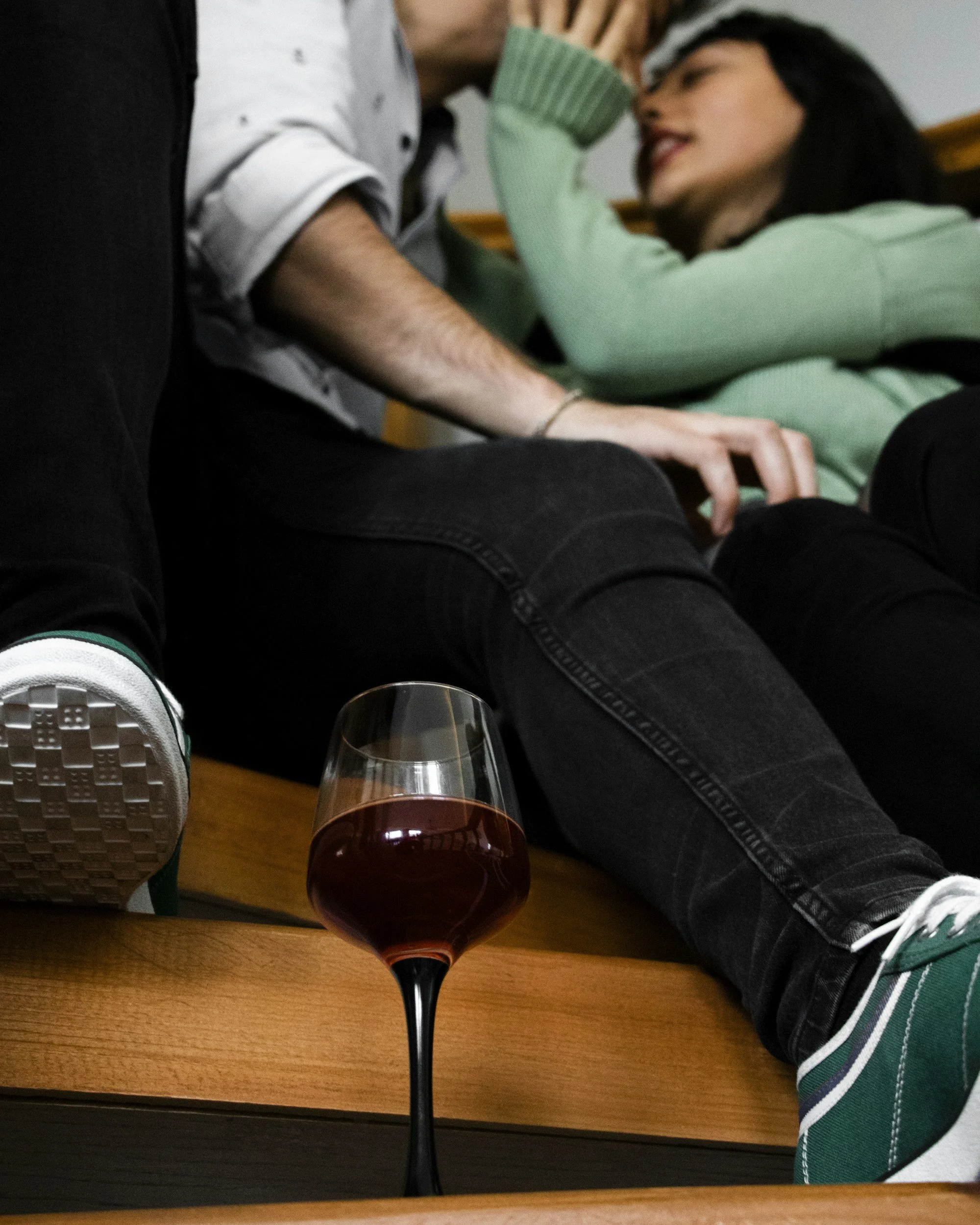 A glass of red wine on a wooden table in front of two people sitting on a bench or couch, one wearing black jeans and sneakers, and the other wearing a green sweater, black pants, and lying back with one arm resting on the other's head.