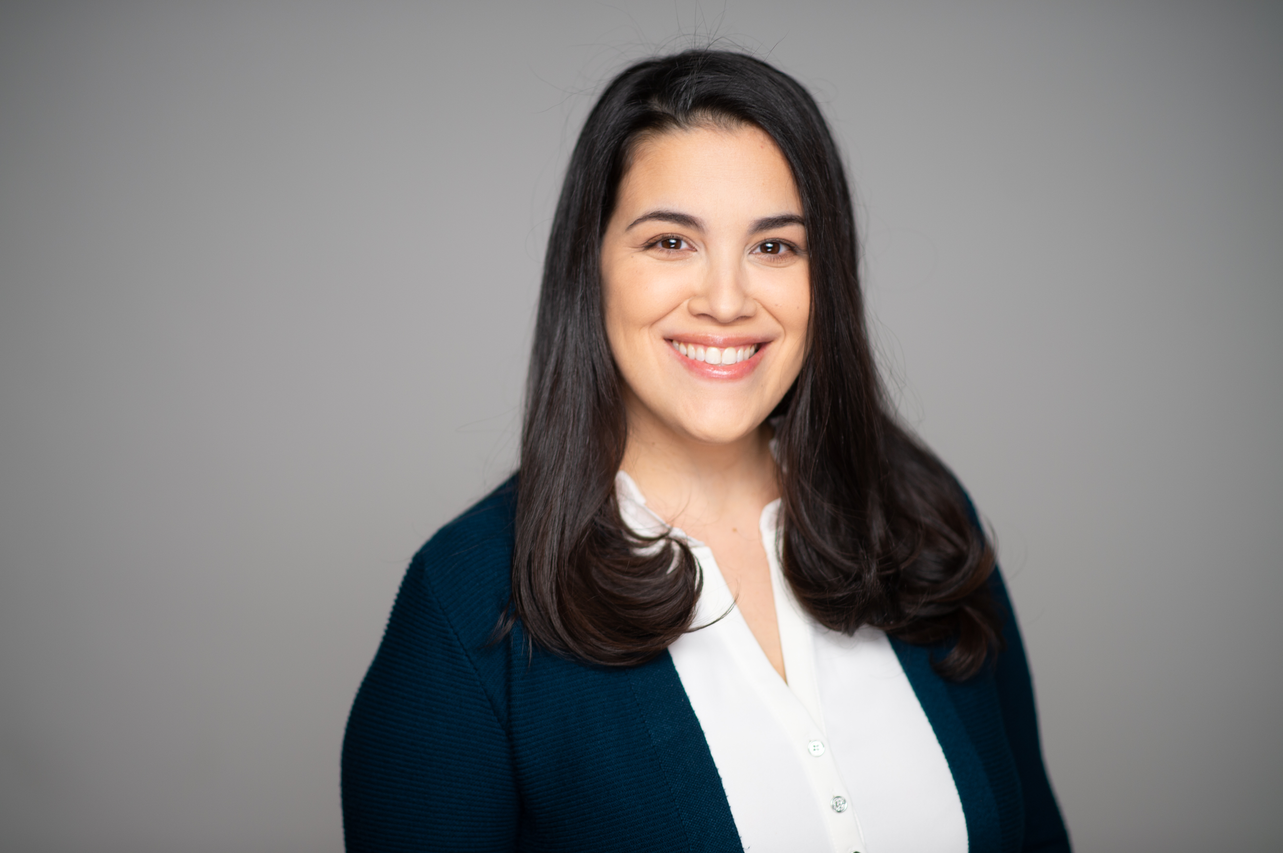 Headshot of a smiling woman with dark brown hair, white shirt, and navy blazer against a neutral gray background.
