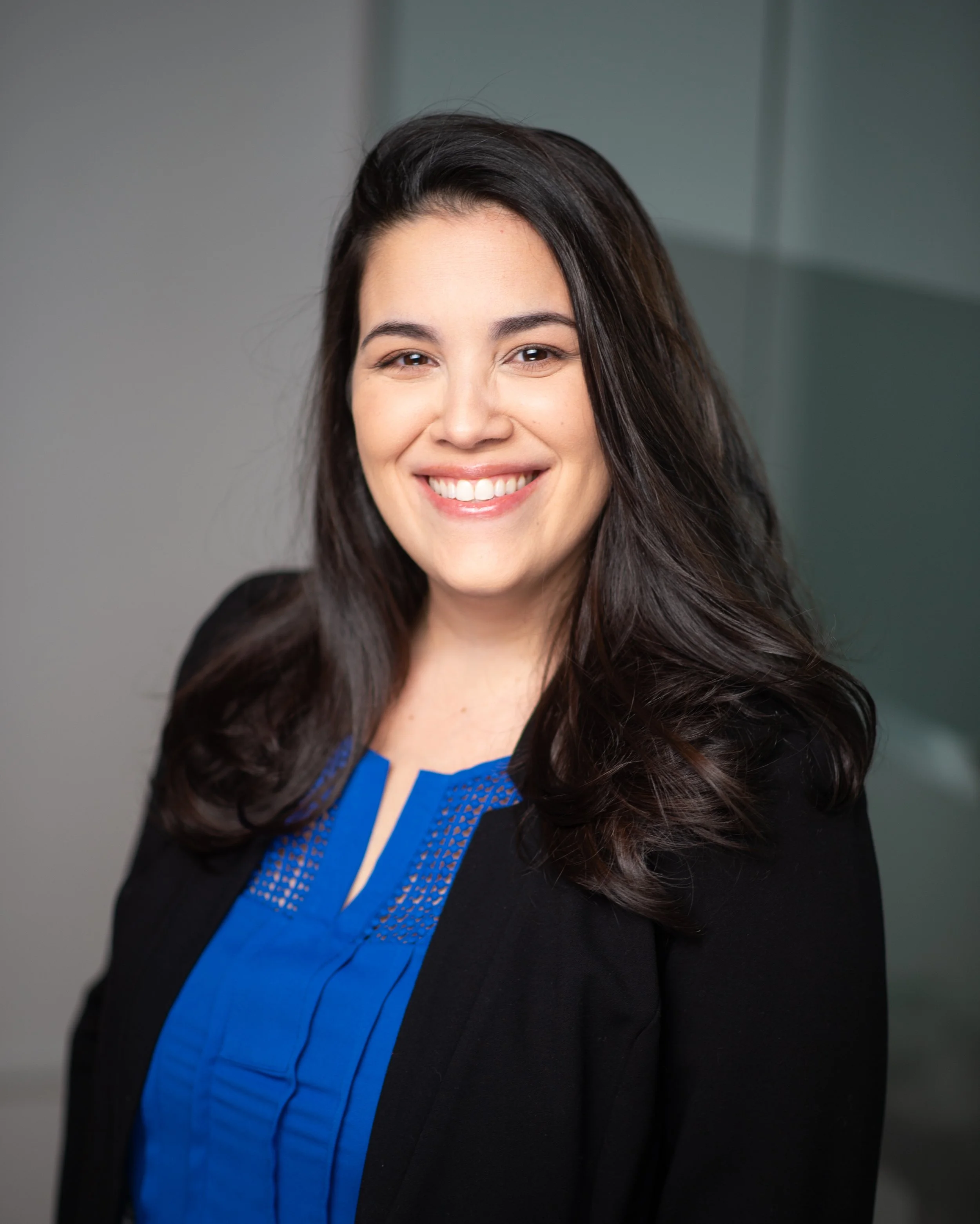 A young woman with long dark hair, smiling, wearing a black blazer and a blue blouse, standing in front of a gray background.