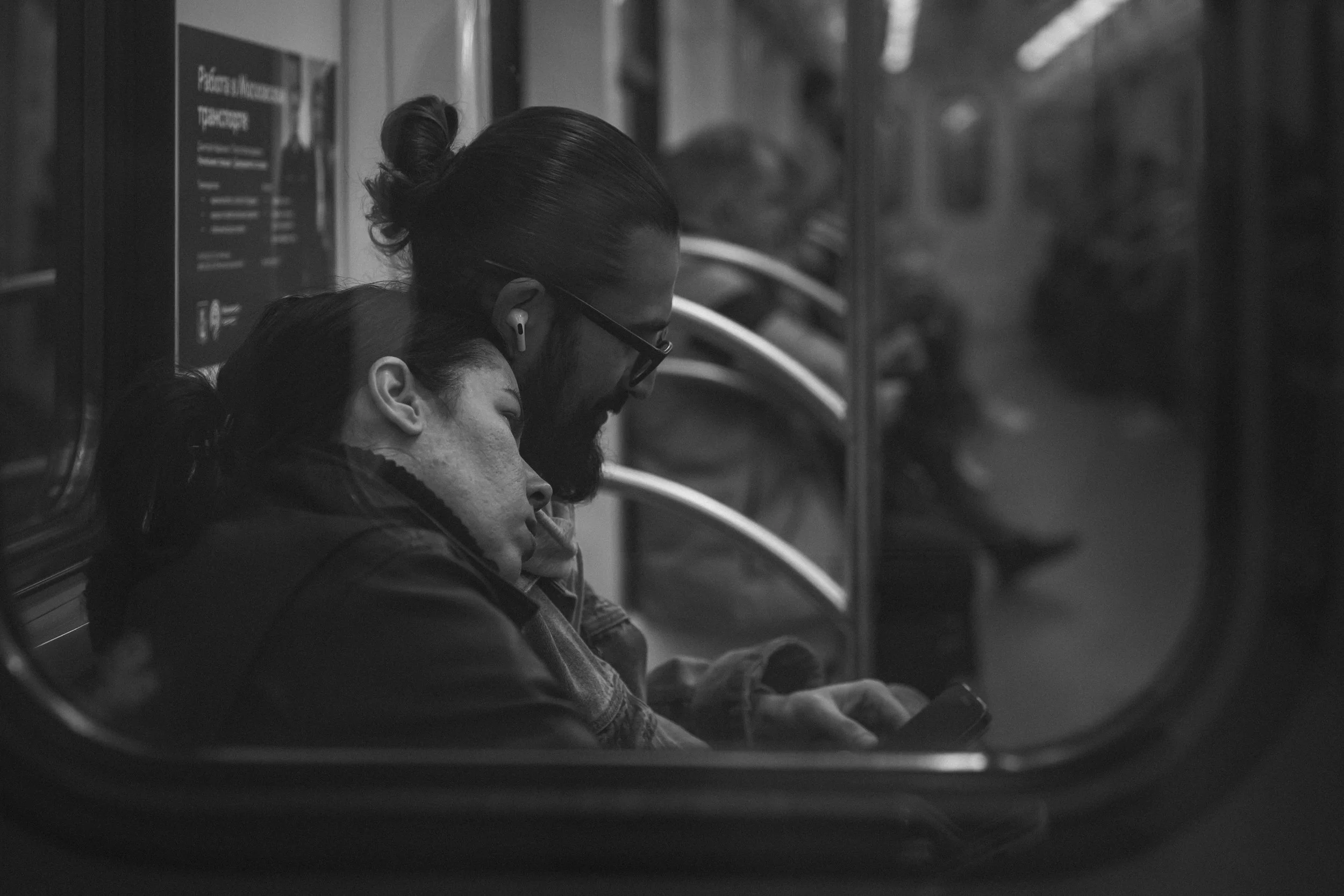 Two people sleeping on a subway train, one wearing wireless earbuds and the other looking at a mobile phone, seen through a subway door window in black and white.