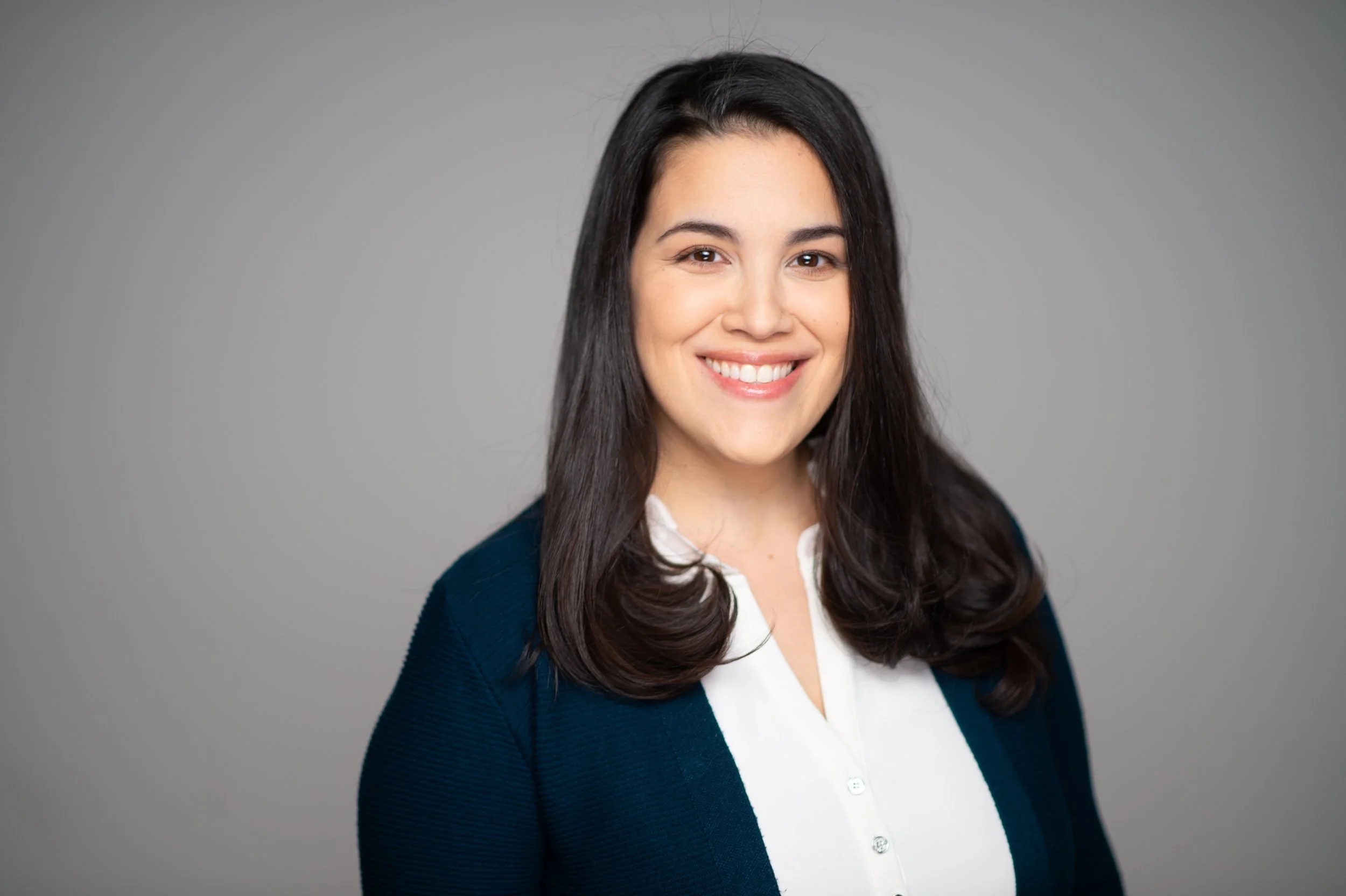 Headshot of a smiling woman with long dark hair, wearing a white blouse and a dark blue blazer, against a gray background.